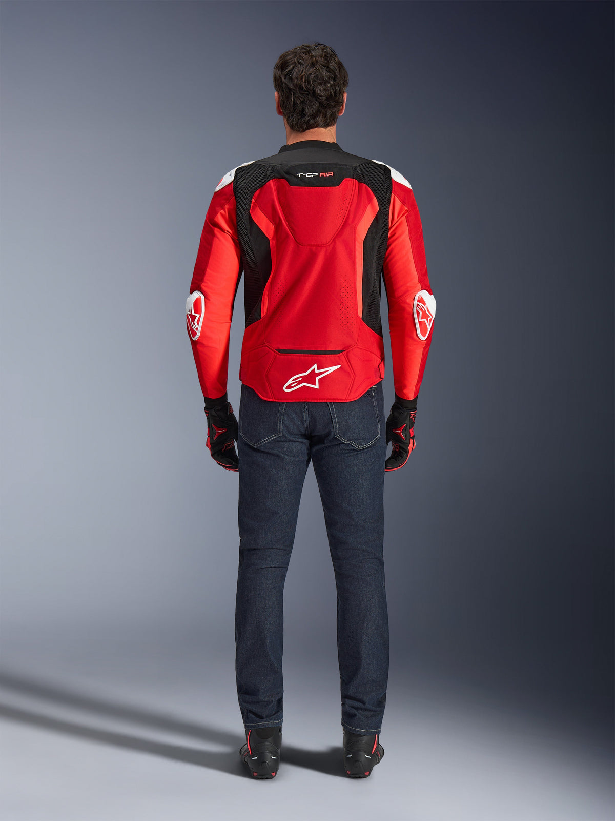 Alpinestars T-GP Air Jacket, Rear view, Bright Red and Black, featuring white logo on the lower back and 'T-GP AIR' branding, worn with dark blue denim jeans and black and red riding gloves, sport-performance motorcycle gear