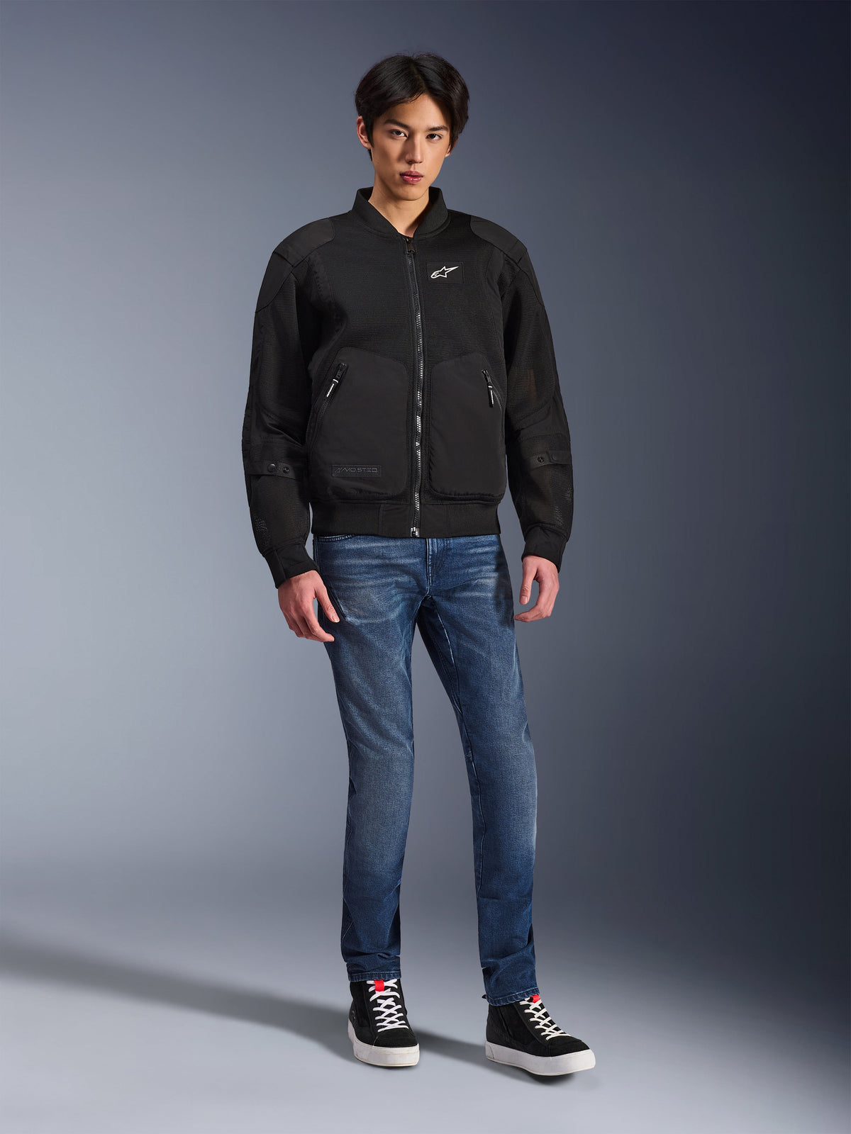 Alpinestars Flight Air Jacke, Schwarz, urban inspirierte Motorradjacke im Bomber-Stil mit 3D-Mesh-Einsätzen und weißem Brustlogo, getragen von einem Model mit blauen Jeans und schwarzen Sneakern, moderne Motorradbekleidung