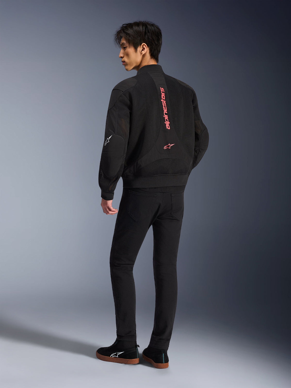 Male model wearing Alpinestars Flight Air Jacket in Black Bright Red, rear view showing vertical red branding on back and 3D mesh construction, paired with black pants and sneakers, urban motorcycle style