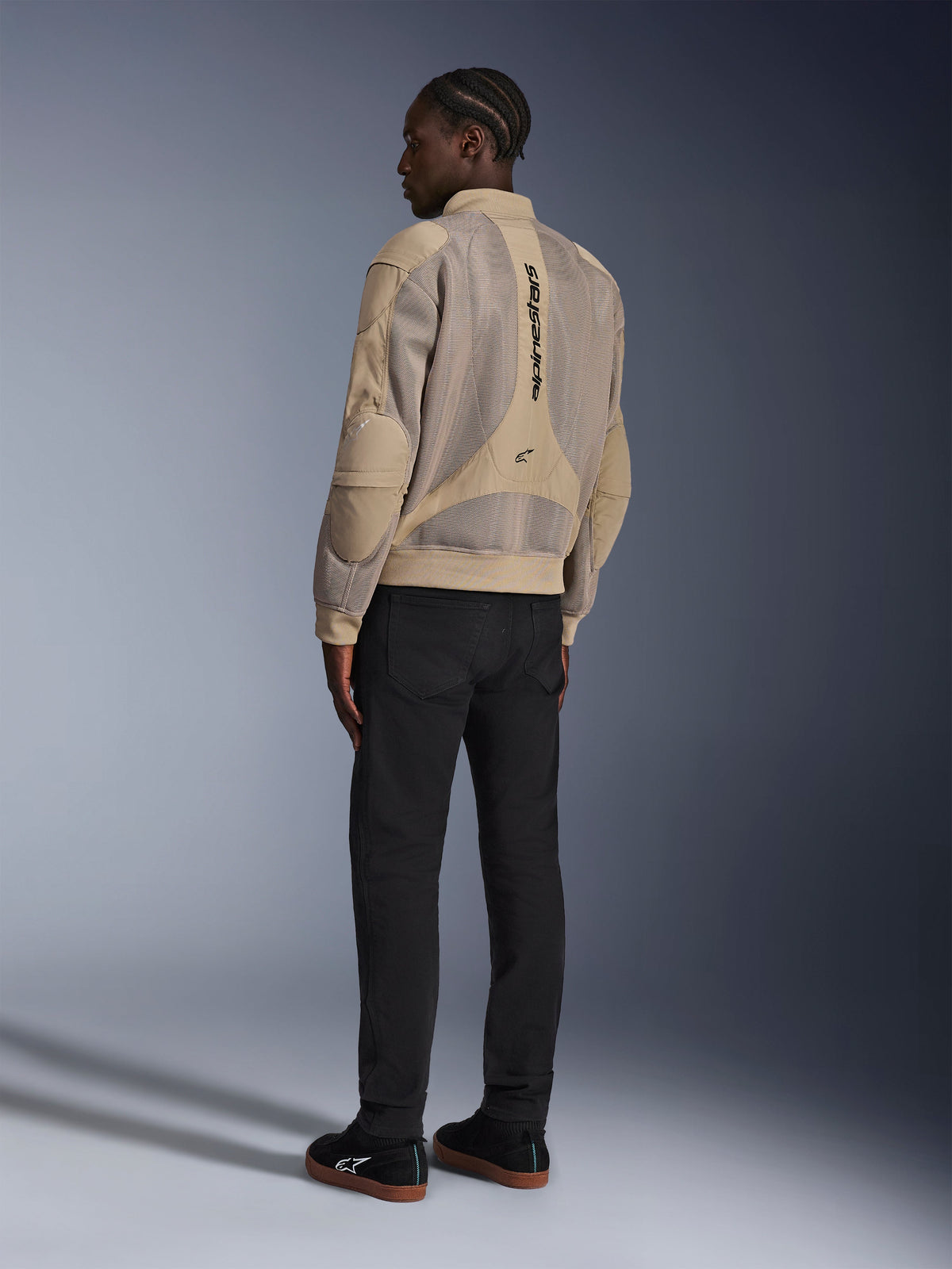 Alpinestars Flight Air Jacke, urbane Textil-Motorradjacke, Braun, Rückansicht mit großen 3D-Mesh-Einsätzen und vertikalem Logo, Bomberjacken-Silhouette mit geripptem Kragen und Saum, kombiniert mit schwarzer Hose und schwarzen Schuhen für einen Street-inspirierten Fahrstil.
