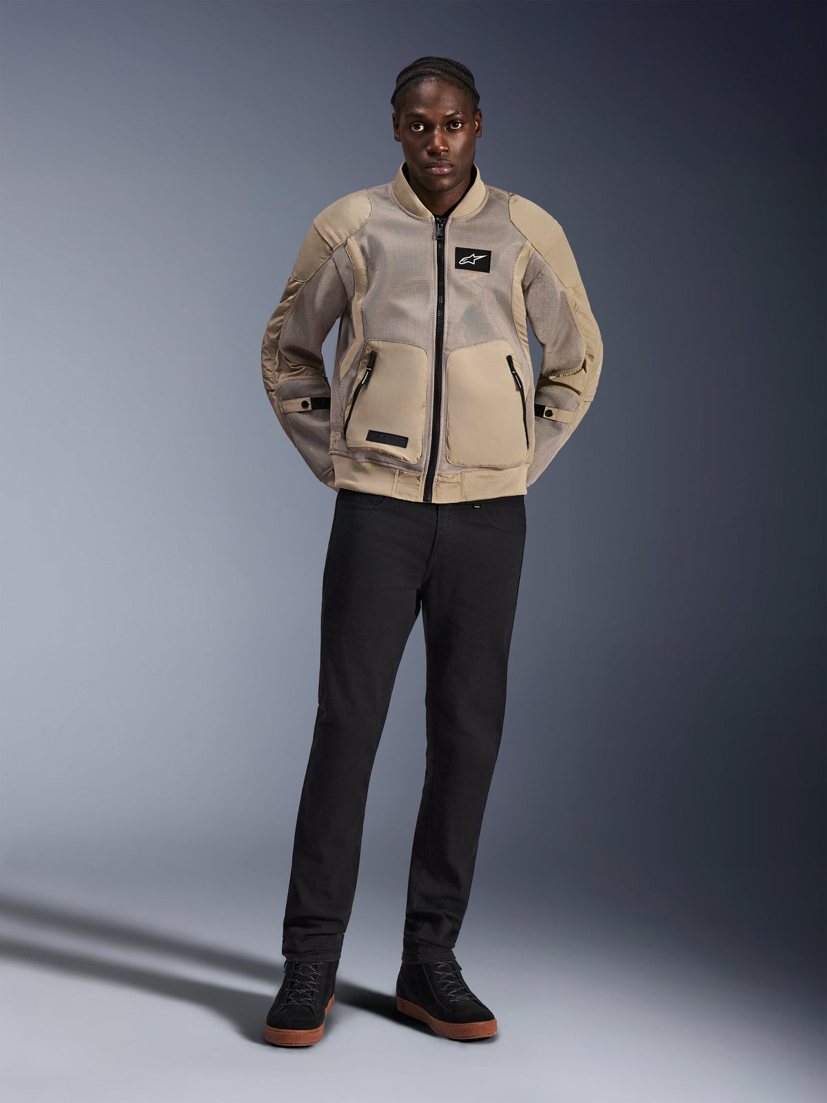 Model trägt die Alpinestars Flight Air Jacke in Braun, urbane Motorradjacke mit erweiterten 3D-Mesh-Einsätzen, Rippstrickkragen und -bündchen im Bomber-Stil, kombiniert mit schwarzer Hose und schwarzen Fahrerschuhen, moderne straßentaugliche Ausrüstung