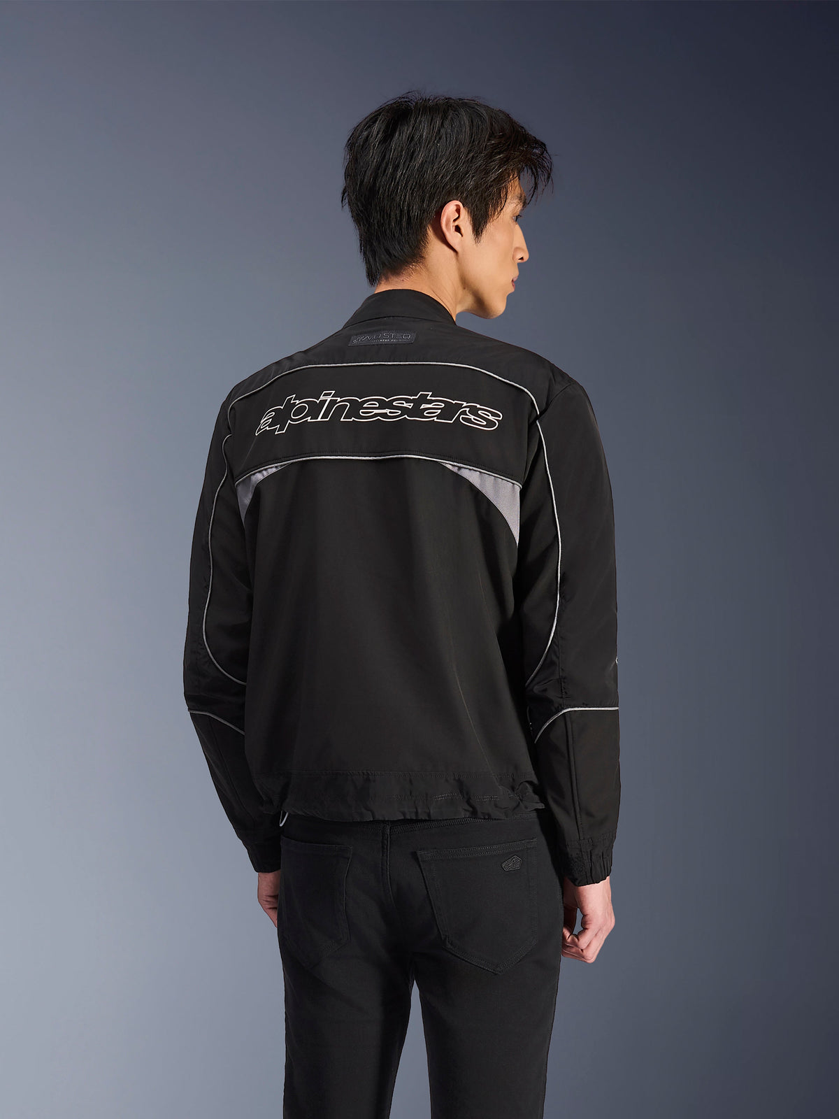 Alpinestars Unite Jacke, Urban-Motorradjacke, Schwarz mit dunkelgrauen Akzenten, Rückansicht mit weißem Alpinestars-Schriftzug über den Schultern, reflektierende Paspeln und Einsätze, minimalistisches Softshell-Streetwear-Design
