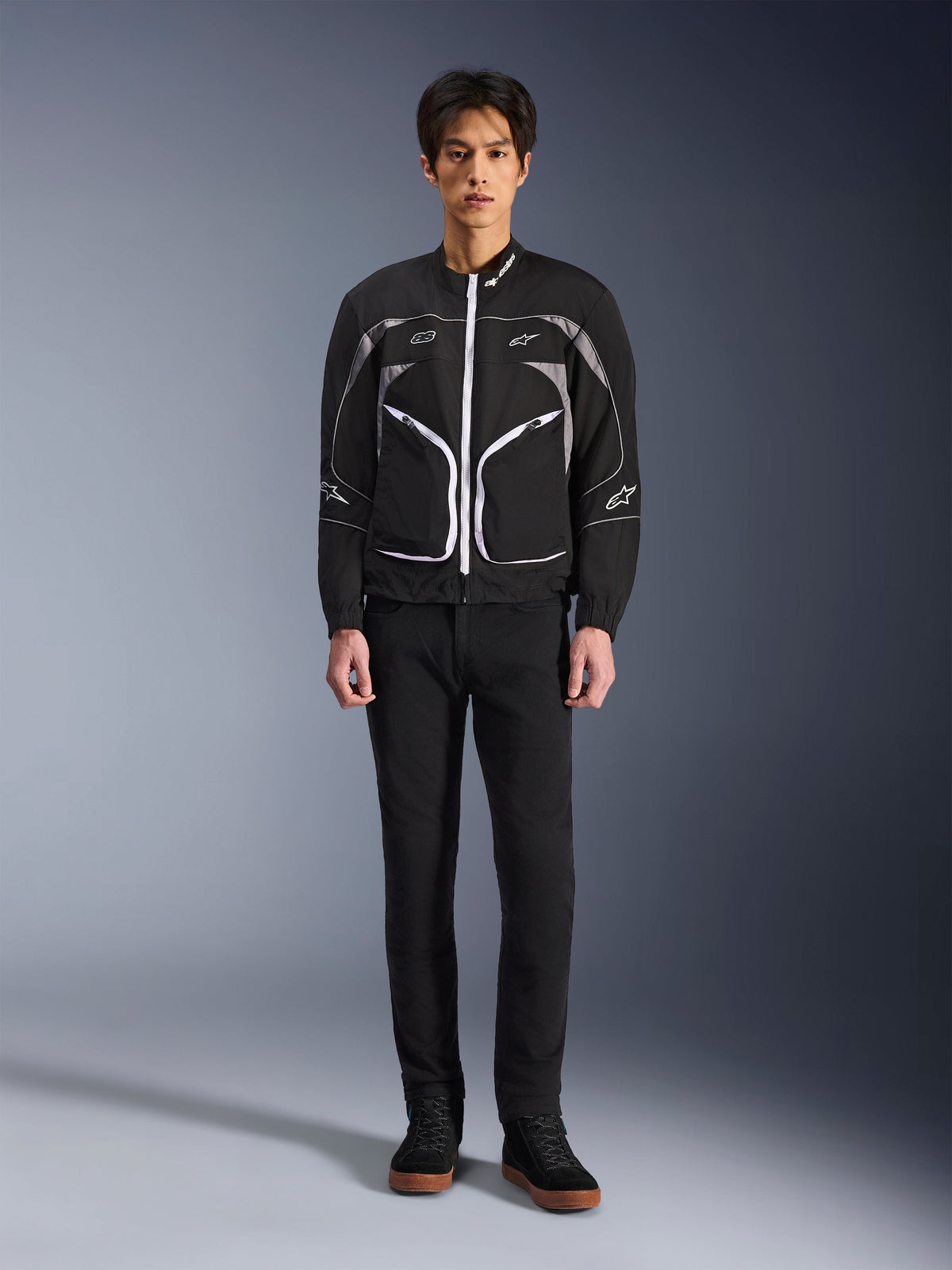 Alpinestars Unite Jacke, Motorradjacke, Schwarz und Dunkelgrau, getragen von einem Model mit schwarzer Hose und schwarzen Stiefeln, weiße Kontrast-Reißverschlüsse, reflektierende Paspeln, minimalistisches urbanes Design