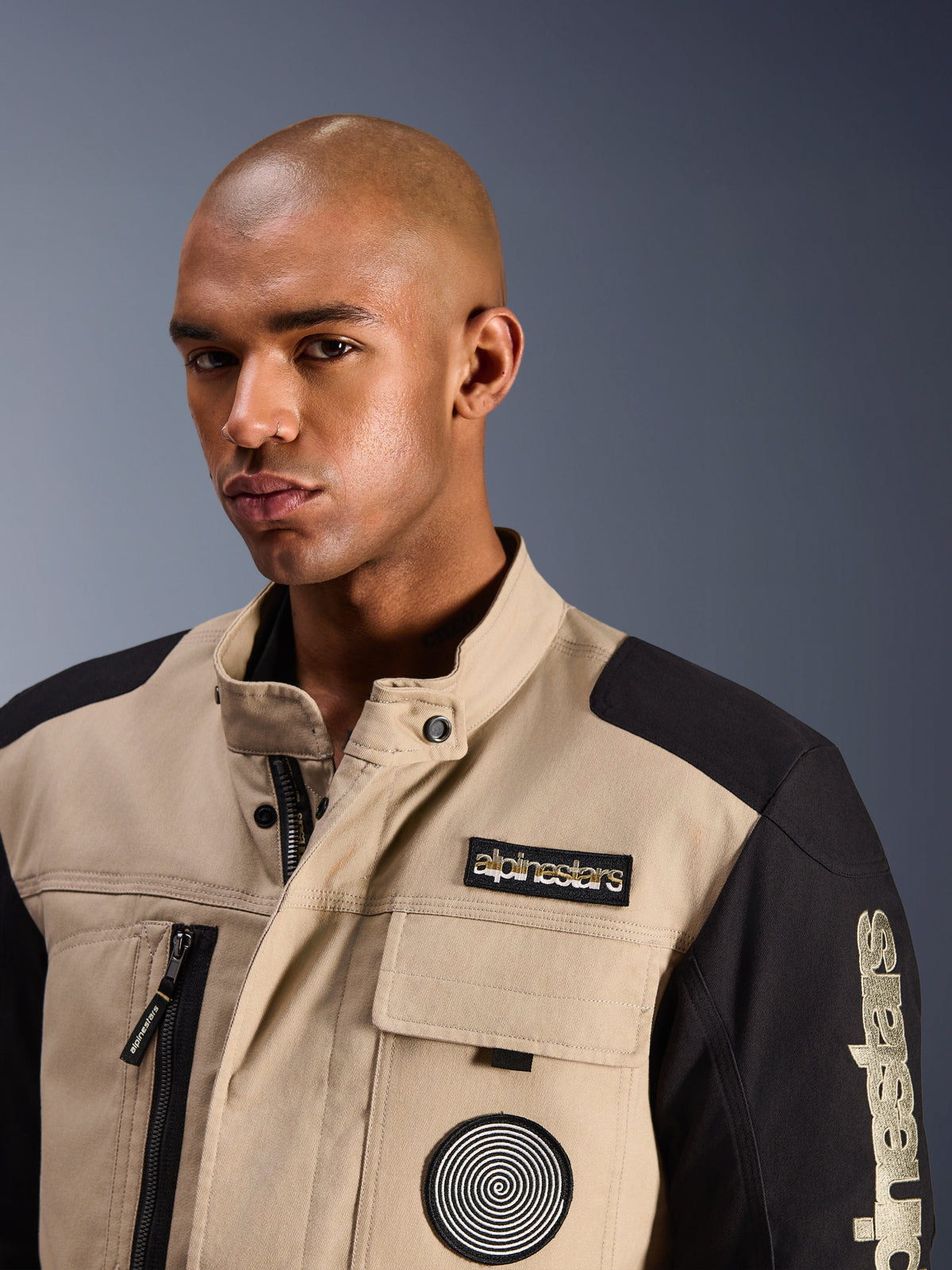 Male model wearing the Alpinestars Dice Jacket in Aluminum Black, urban motorcycle jacket with tan and black color-blocked panels, featuring an embroidered Alpinestars chest patch and spiral logo, rugged workwear-inspired design with a high collar.