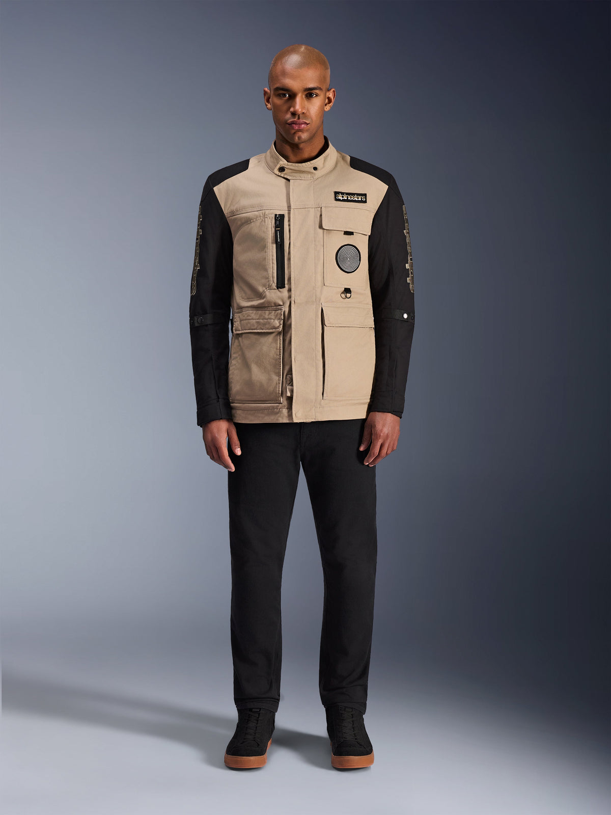 Alpinestars Dice Jacket, urban motorcycle jacket, aluminum tan body with black sleeves, workwear-inspired silhouette with four front utility pockets, worn with black pants and black high-top sneakers, rugged street-ready riding gear
