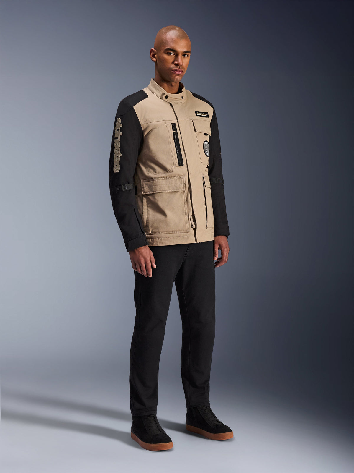 Alpinestars Dice Jacket, Urban Motorcycle Jacket, Aluminum Black and Tan, two-tone design with black sleeves and a khaki torso, worn by a model with black pants and black sneakers, workwear-inspired aesthetic with utility pockets and embroidered branding on the sleeve.
