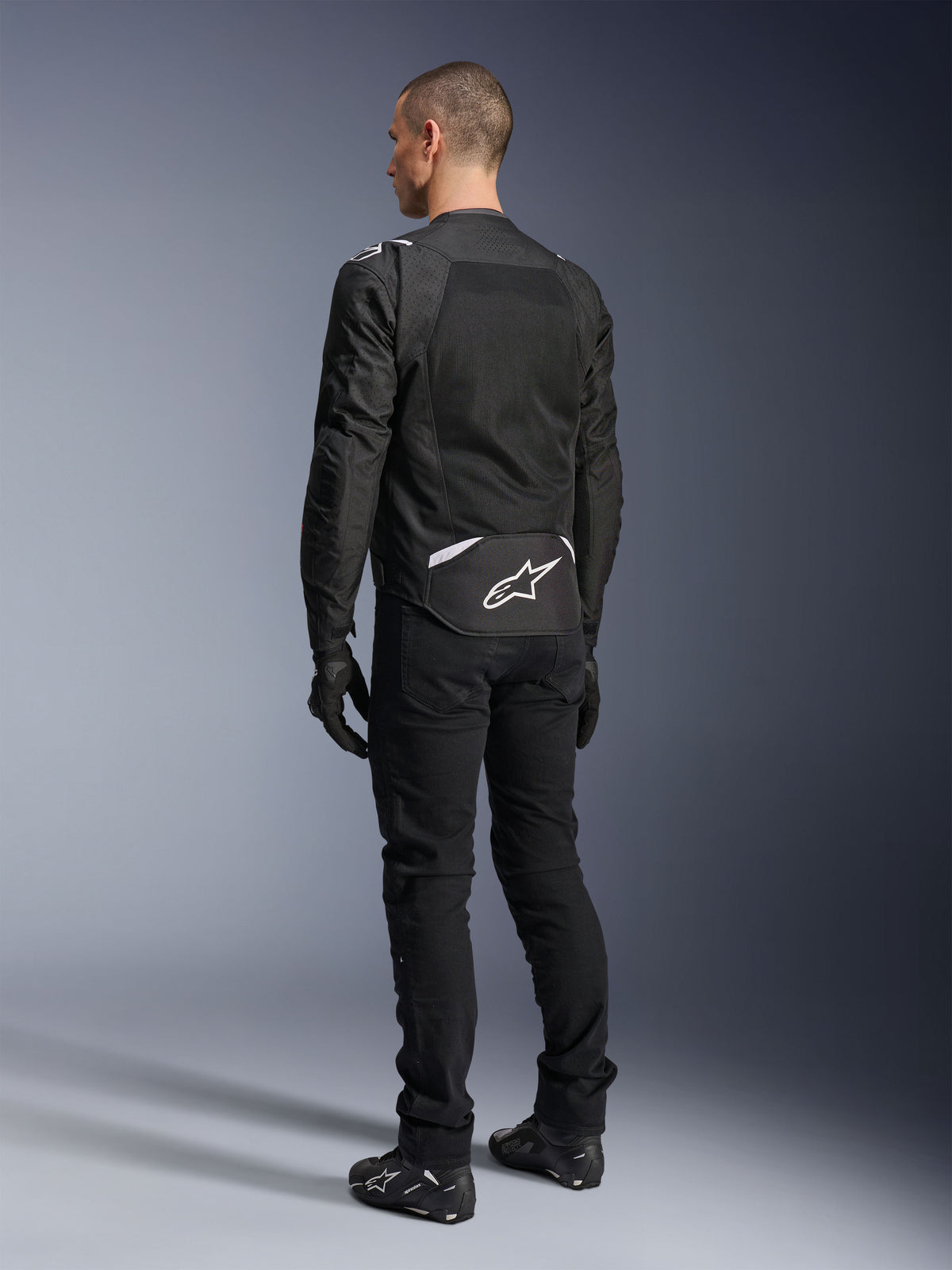 Alpinestars T-Sps Air V2 Jacke, Textil-Motorradjacke, Schwarz, Rückansicht mit großem Mesh-Belüftungspanel und weißem Alpinestars-Logo am unteren Rücken, kombiniert mit schwarzer Motorradhose und Handschuhen für einen sportlichen urbanen Look.