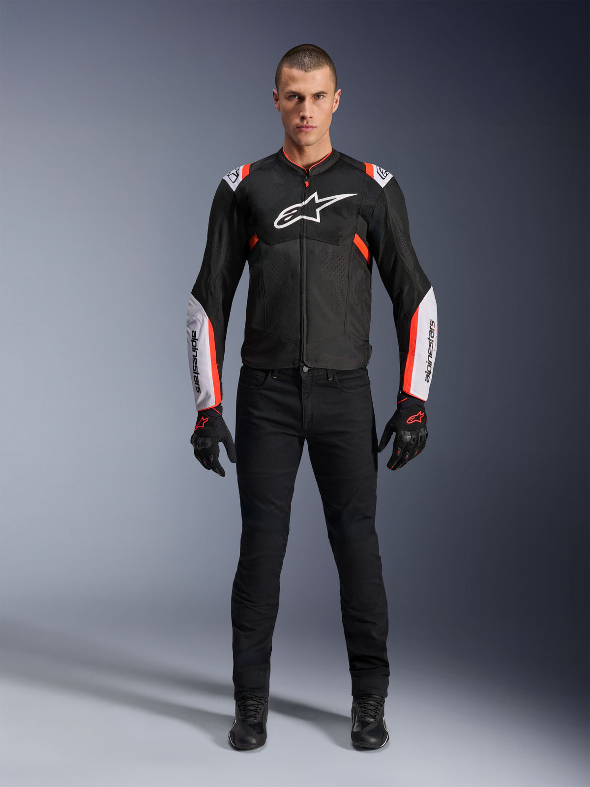 Männliches Model in der Alpinestars T-Sps Air V2 Jacke in Schwarz Weiß Rot Fluo, mit großen Tech-Mesh-Einsätzen und markantem weißem Branding auf Brust und Ärmeln, kombiniert mit schwarzer technischer Motorradhose und Handschuhen, urbane Sport-Motorradausrüstung