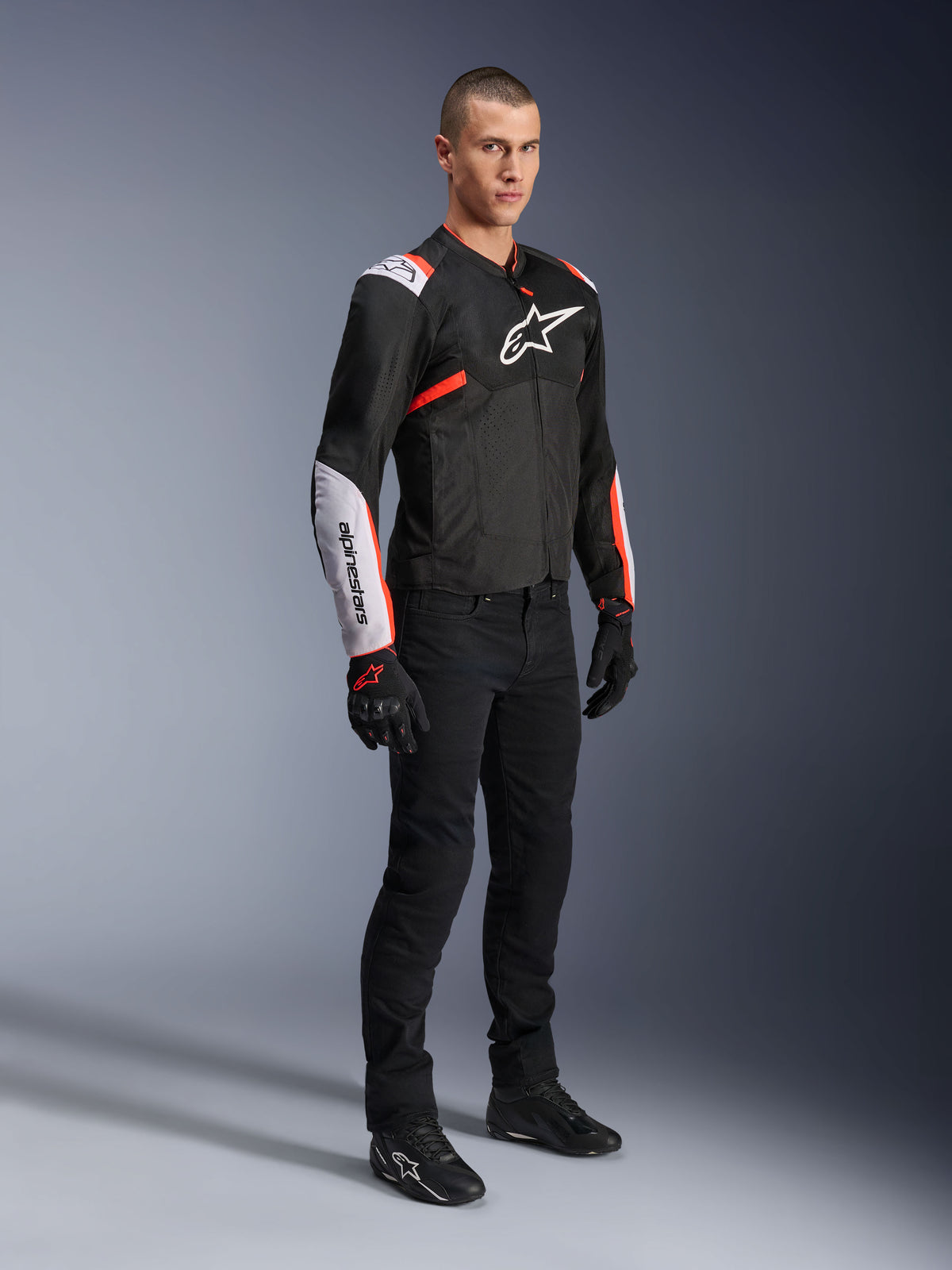 Alpinestars T-Sps Air V2 Jacke, Schwarz Weiß Rot Fluo, getragen von einem männlichen Model mit schwarzer Motorradhose und Handschuhen, Textil-Motorradjacke mit großen Mesh-Einsätzen, weißen Schulter- und Unterarmbereichen, markantes Alpinestars-Logo auf der Brust.