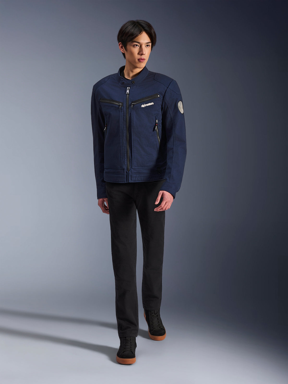 Männliches Model in Alpinestars Bruiser Jacke in Dark Blue, urbane Textil-Motorradjacke, kombiniert mit schwarzer Hose und schwarzen Sneakern, Workwear-inspirierte Ästhetik mit Reißverschlusstaschen und rundem Ärmel-Patch, zeitgemäße Streetwear für das Motorradfahren.