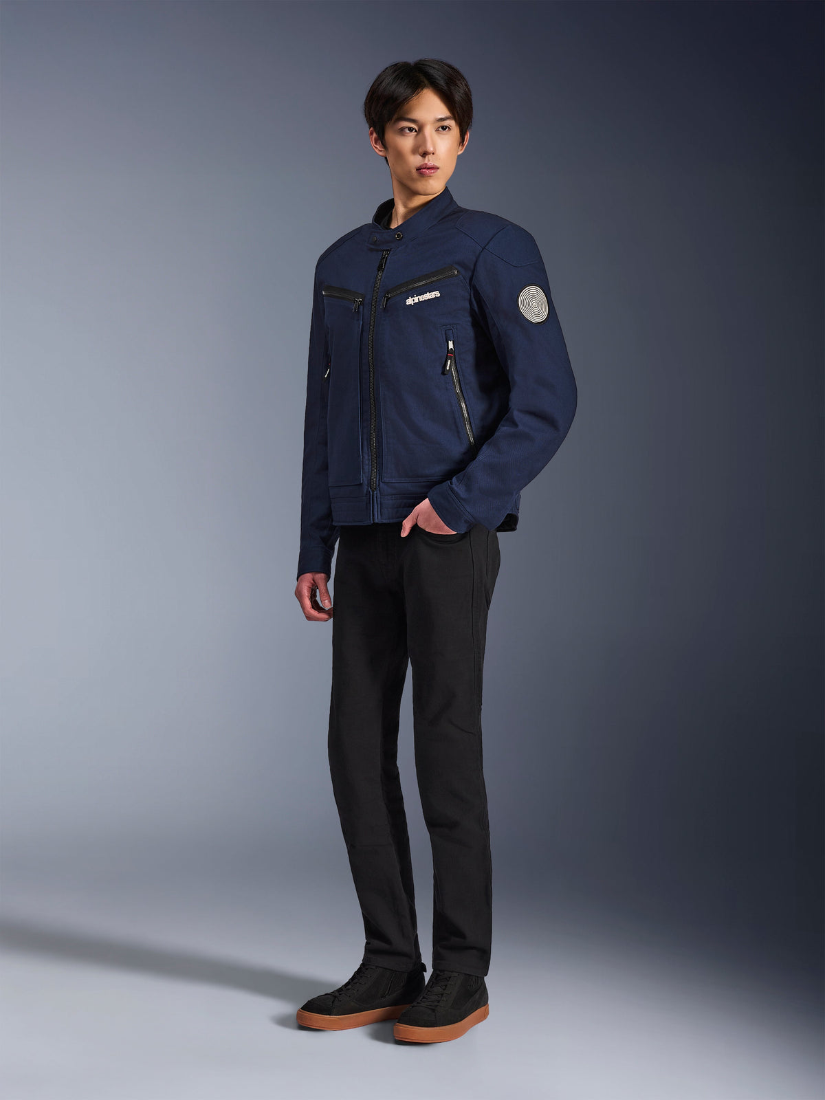 Alpinestars Bruiser Jacke in Dunkelblau, urbane Textil-Motorradjacke mit von Workwear inspiriertem Design, mit Reißverschlusstaschen an der Brust und Oscar Spiral-Ärmel-Patch, getragen mit schwarzer Hose und lässigen Sneakern.