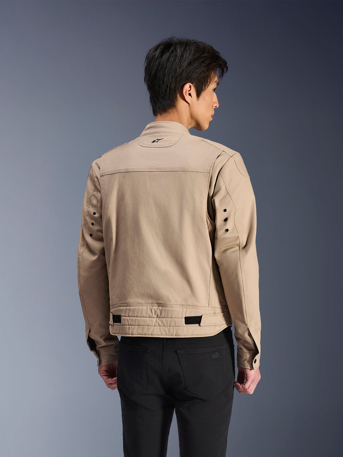 Alpinestars Bruiser Jacke, braune urbane Motorradjacke, Rückansicht mit plissierten Schultern und verstellbaren Taillenriemen, getragen mit schwarzer Hose, moderne, von Arbeitskleidung inspirierte Fahrausrüstung