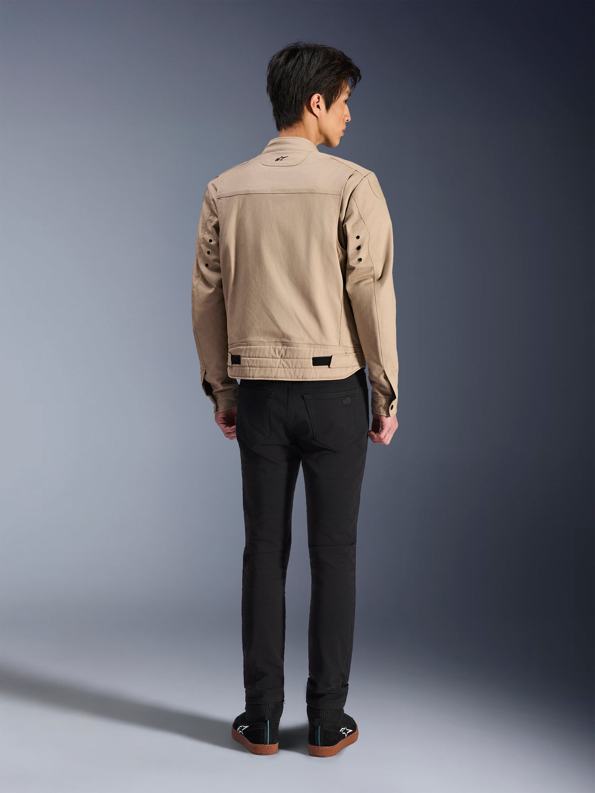 Rückansicht eines Modells, das die Alpinestars Bruiser Jacke in Braun trägt, eine urbane Motorradjacke mit von Workwear inspiriertem Design, mit Metallösen am Rücken und Taillenverstellern, kombiniert mit schwarzer Hose und Sneakern, moderner Street-Riding-Stil.