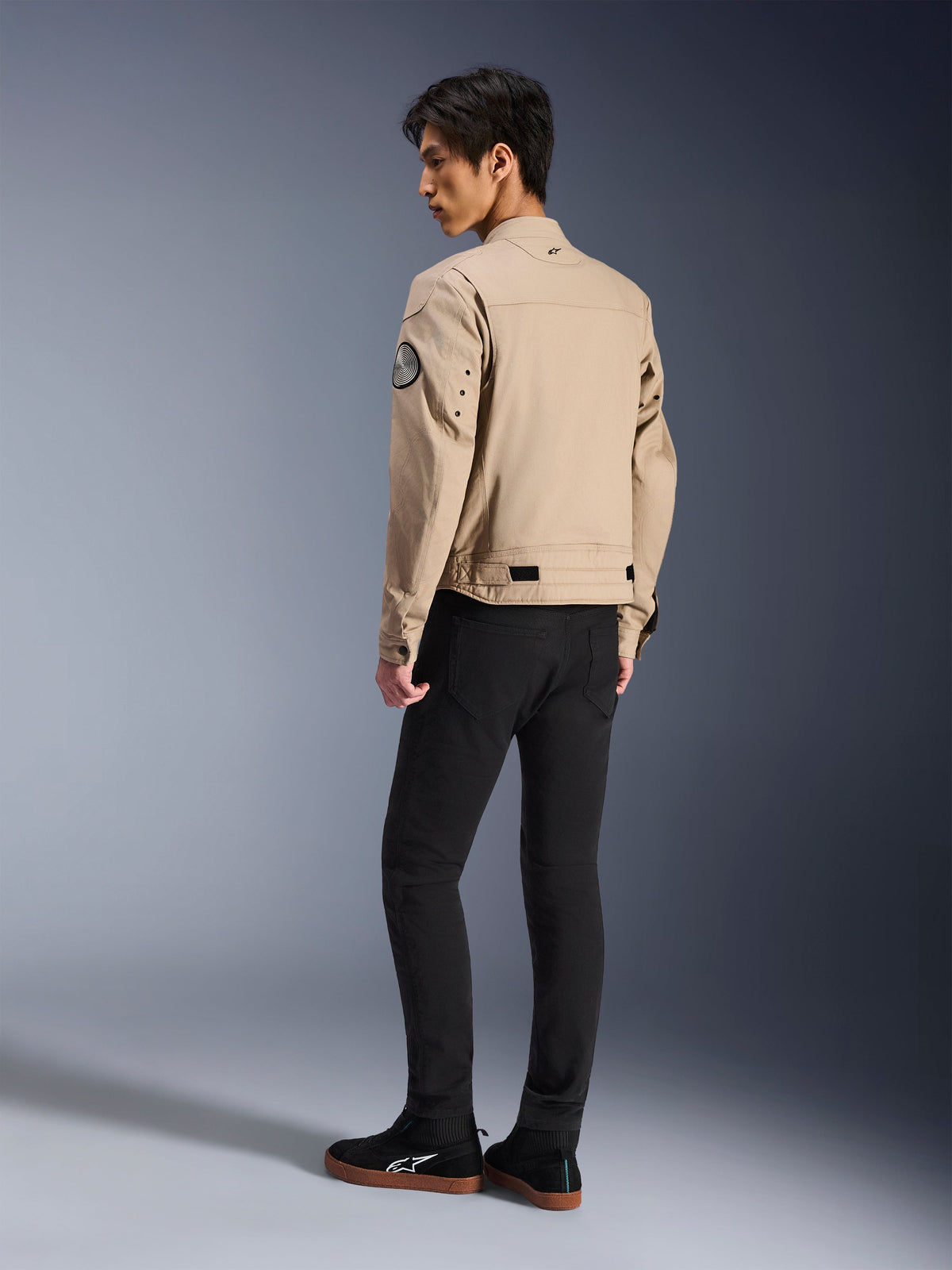 Männliches Model von hinten in der Alpinestars Bruiser Jacke in Tan, urbane moderne Motorradjacke im Workwear-Stil, mit rundem Ärmel-Patch, Taillenverstellriemen und vorgeformten Schultern, kombiniert mit schwarzer Hose und schwarzen Sneakern.