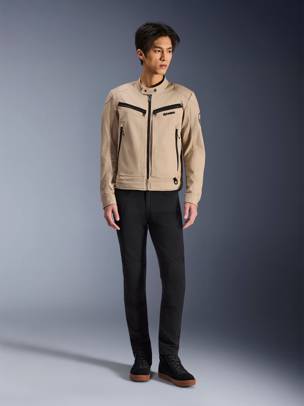 Alpinestars Bruiser Jacke, urbane Motorradjacke in Aluminum, getragen mit schwarzer Hose und schwarzen Sneakern, mit schwarzen Reißverschlusstaschen und einem Druckknopfkragen, zeitgenössisches, von Workwear inspiriertes Design für urbanes Fahren