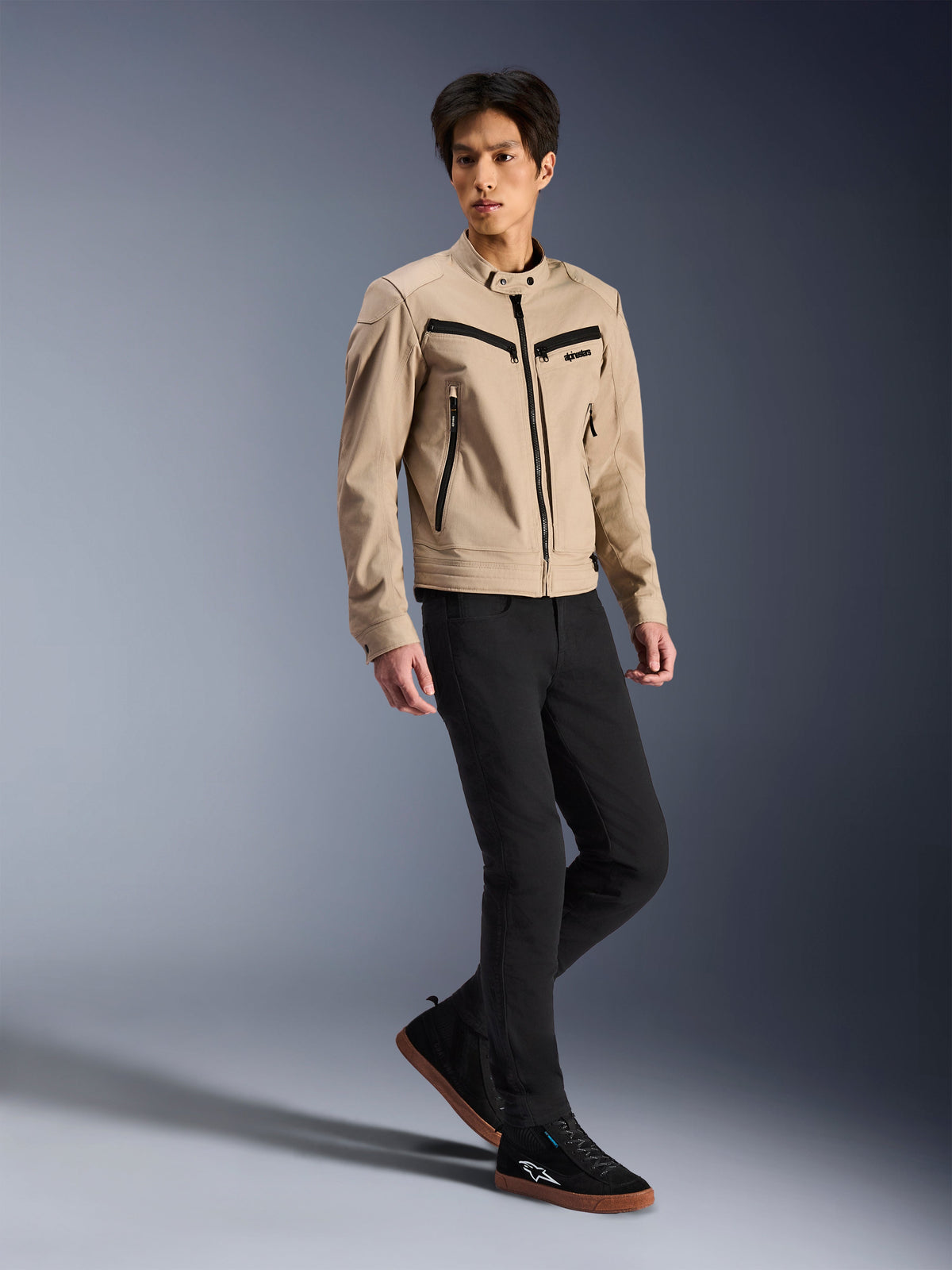 Alpinestars Bruiser Jacke, urbane Motorradjacke, Tan, getragen von einem männlichen Model mit schwarzer Slim-Fit-Hose und schwarzen Sneakern, von Workwear inspirierte Textilkonstruktion mit schwarzen Reißverschluss-Akzenten, zeitgenössischer urbaner Streetstyle.