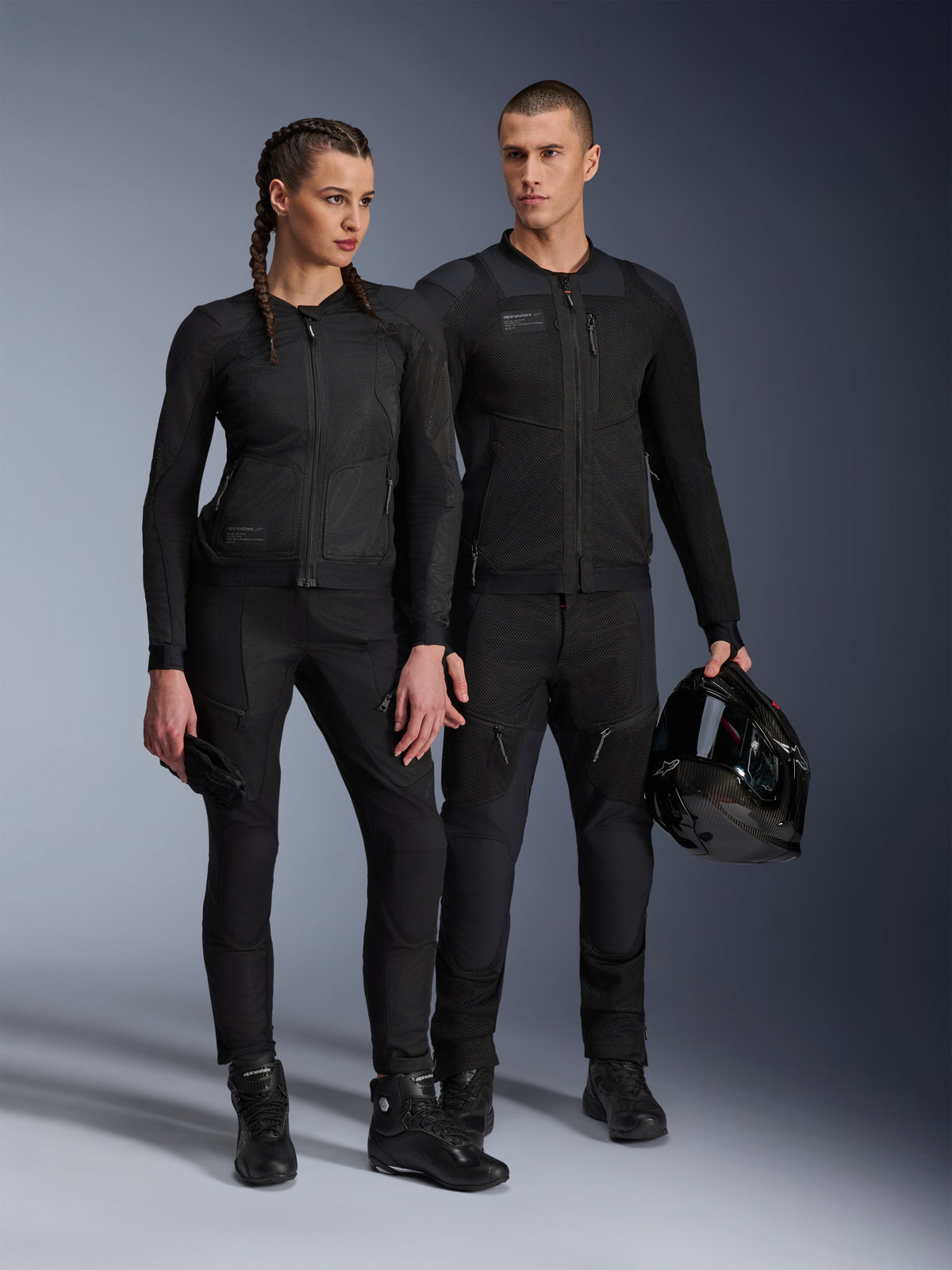 Alpinestars Stella Troop-Air Jacke, Mann und Frau tragen passende, komplett schwarze technische Motorradausrüstung, mit Mesh-Konstruktion für optimale Belüftung, schwarze Hosen und Stiefel, der Mann hält einen schwarzen Helm, urbane Motorradbekleidung