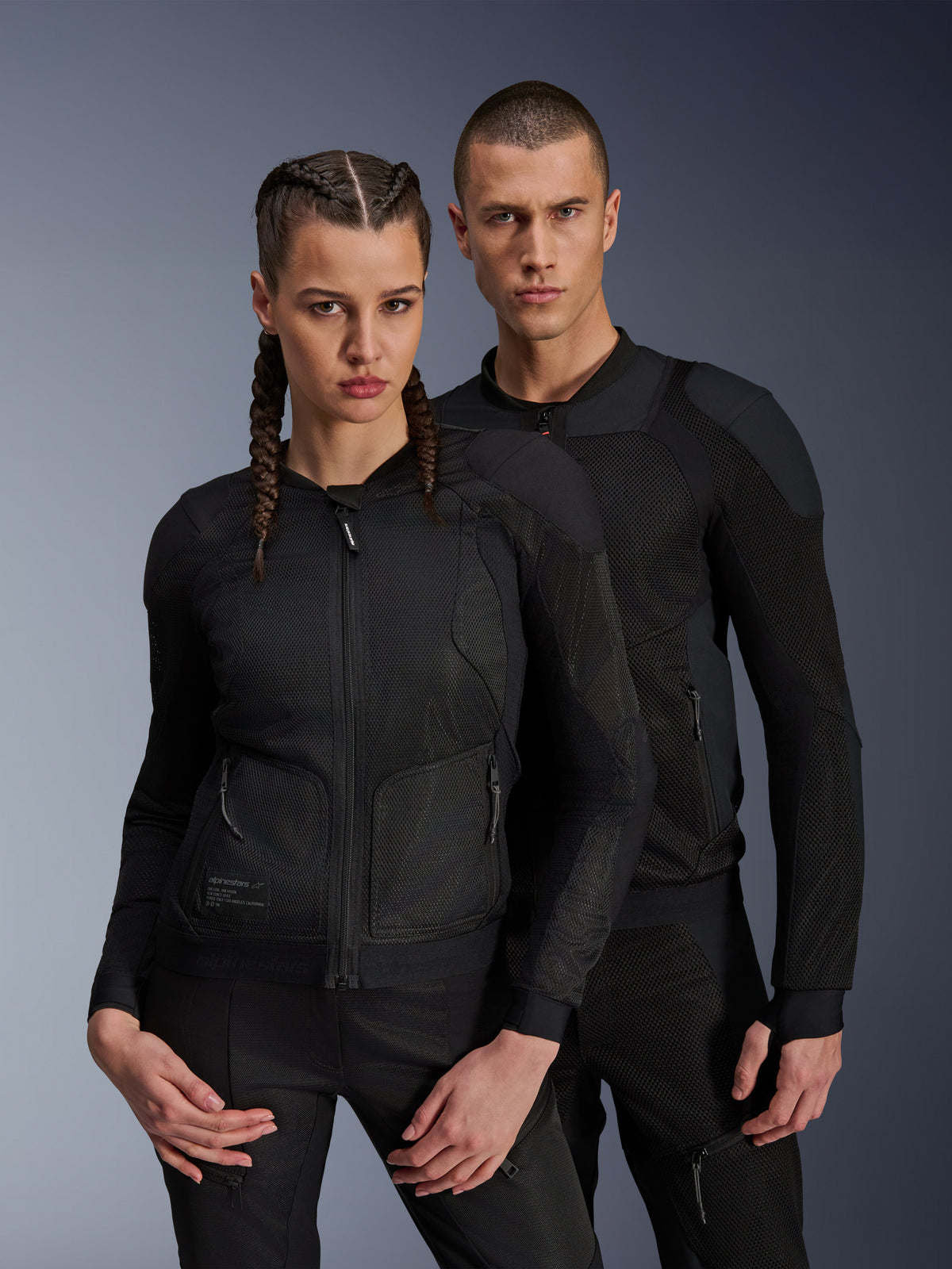 Mann und Frau in Alpinestars Troop-Air und Stella Troop-Air Jacken, schwarze technische Mesh-Motorradjacken mit 2D-Tech-Mesh-Struktur zur Belüftung und verstärkten Einsätzen, moderne urbane Fahrbekleidung.
