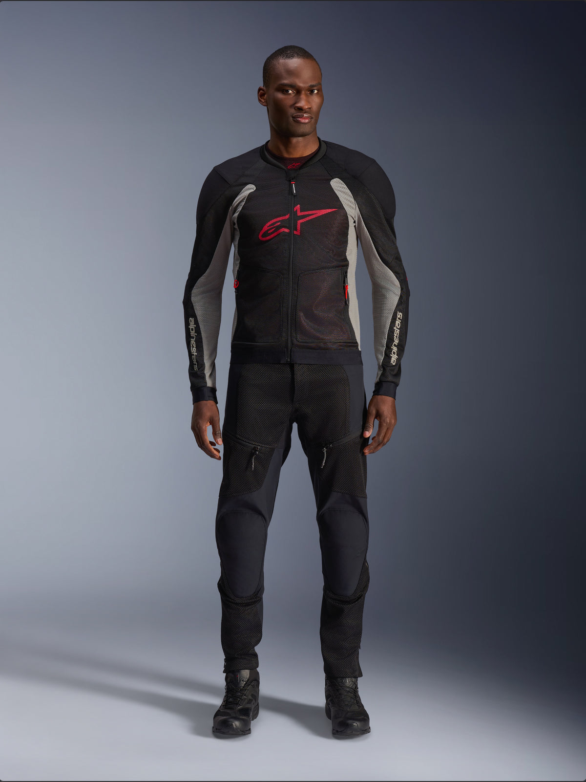 Männliches Model in der Alpinestars Troop-Air Jacke in Black Ash Gray und Bright Red, technische Mesh-Motorradjacke mit markantem roten Brustlogo, kombiniert mit schwarzer Motorradhose und Stiefeln, urbane Motorradbekleidung