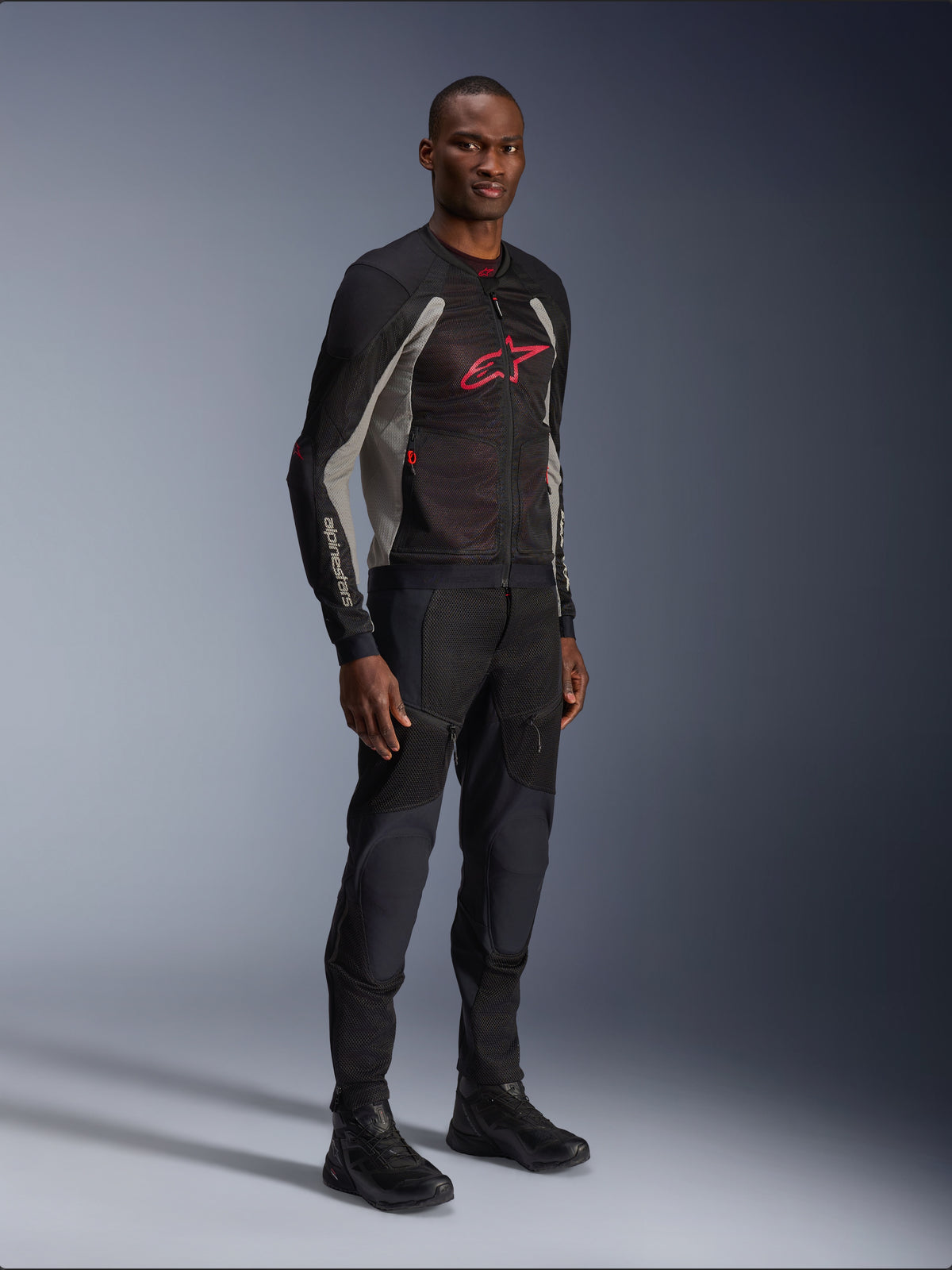 Alpinestars Troop-Air Jacke, technische Mesh-Motorradjacke in Schwarz, Ash Gray und Bright Red, getragen von einem Modell mit passender schwarzer Hose und Stiefeln, mit rotem Alpinestars-Logo auf der Brust, urbane zeitgemäße Motorradausrüstung.