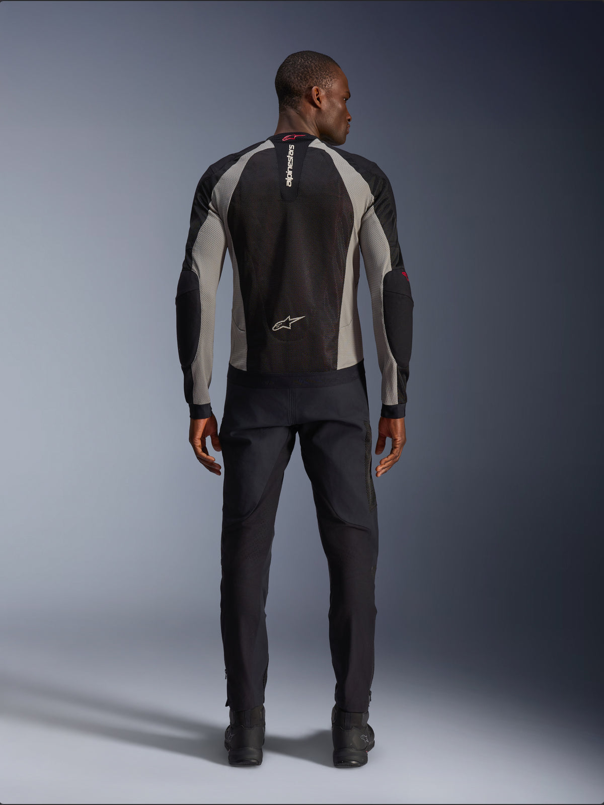 Alpinestars Troop-Air Jacke, Motorradjacke in Black Ash Gray und Bright Red, Rückansicht an einem männlichen Model, leichte Mesh-Konstruktion für Belüftung, Alpinestars-Branding am Kragen und im unteren Rückenbereich, kombiniert mit einer schwarzen Fahrhose.