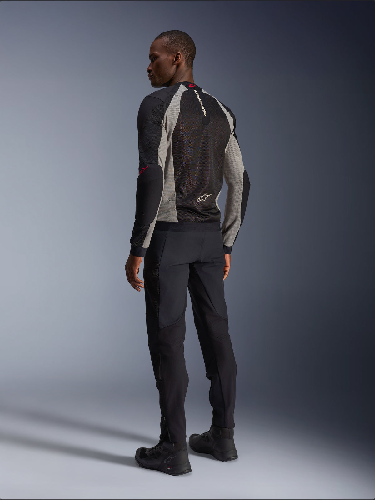 Model trägt Alpinestars Troop-Air Jacke in der Rückansicht, Schwarz, Aschgrau mit leuchtend roten Akzenten, technische Mesh-Konstruktion zur Belüftung, kombiniert mit schwarzer Motorradhose und Fahrstiefeln, urbaner zeitgenössischer Stil