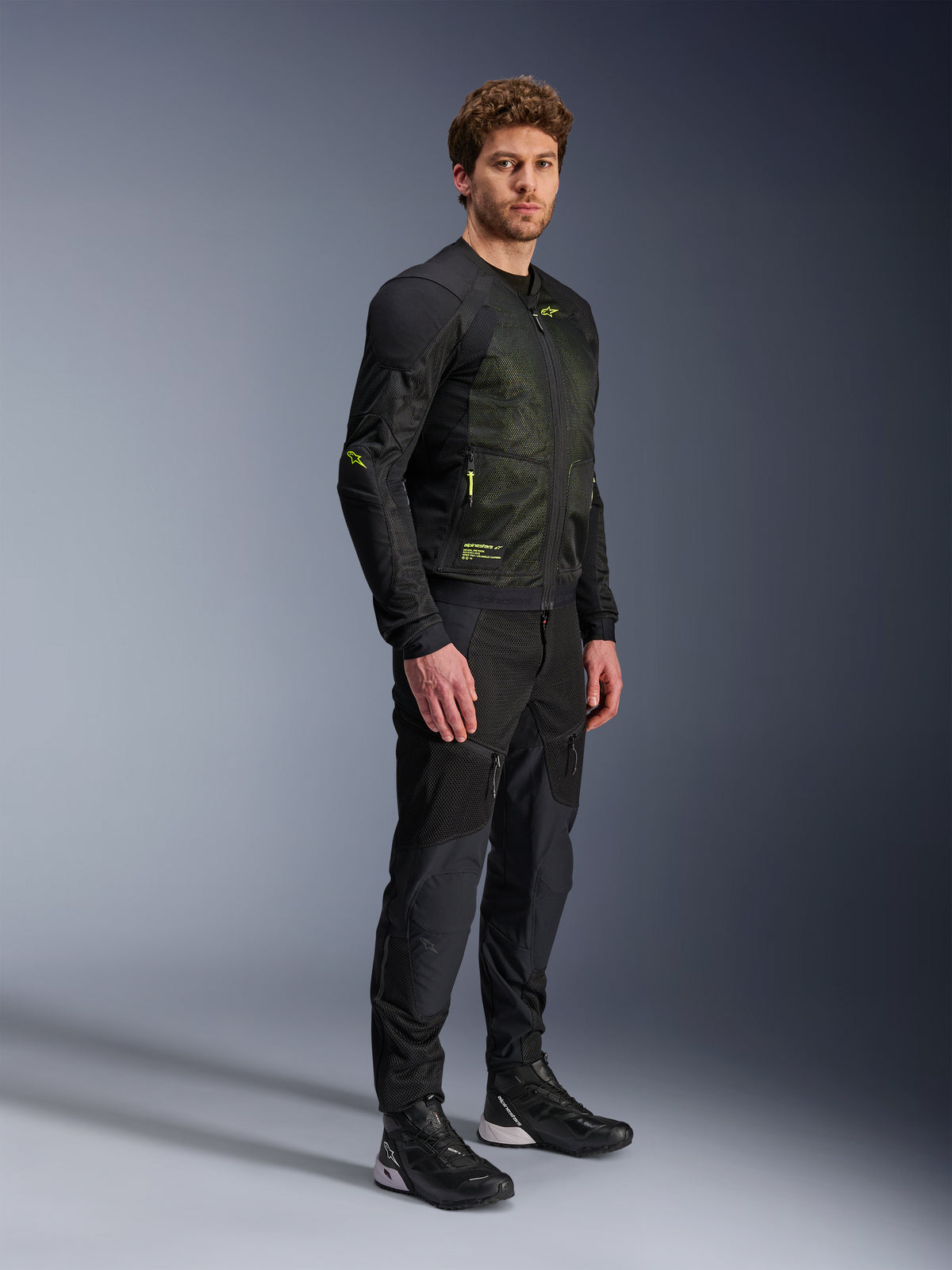 Alpinestars Troop-Air Jacke, Motorrad-Mesh-Jacke, Schwarz und Gelb Fluo, getragen von einem Modell mit passender schwarzer Hose und Motorradschuhen, verfügt über eine Mesh-Konstruktion für hohen Luftstrom mit gelben fluoreszierenden Akzenten an Brust und Ärmeln, leichtes technisches Design für Straßenfahrten