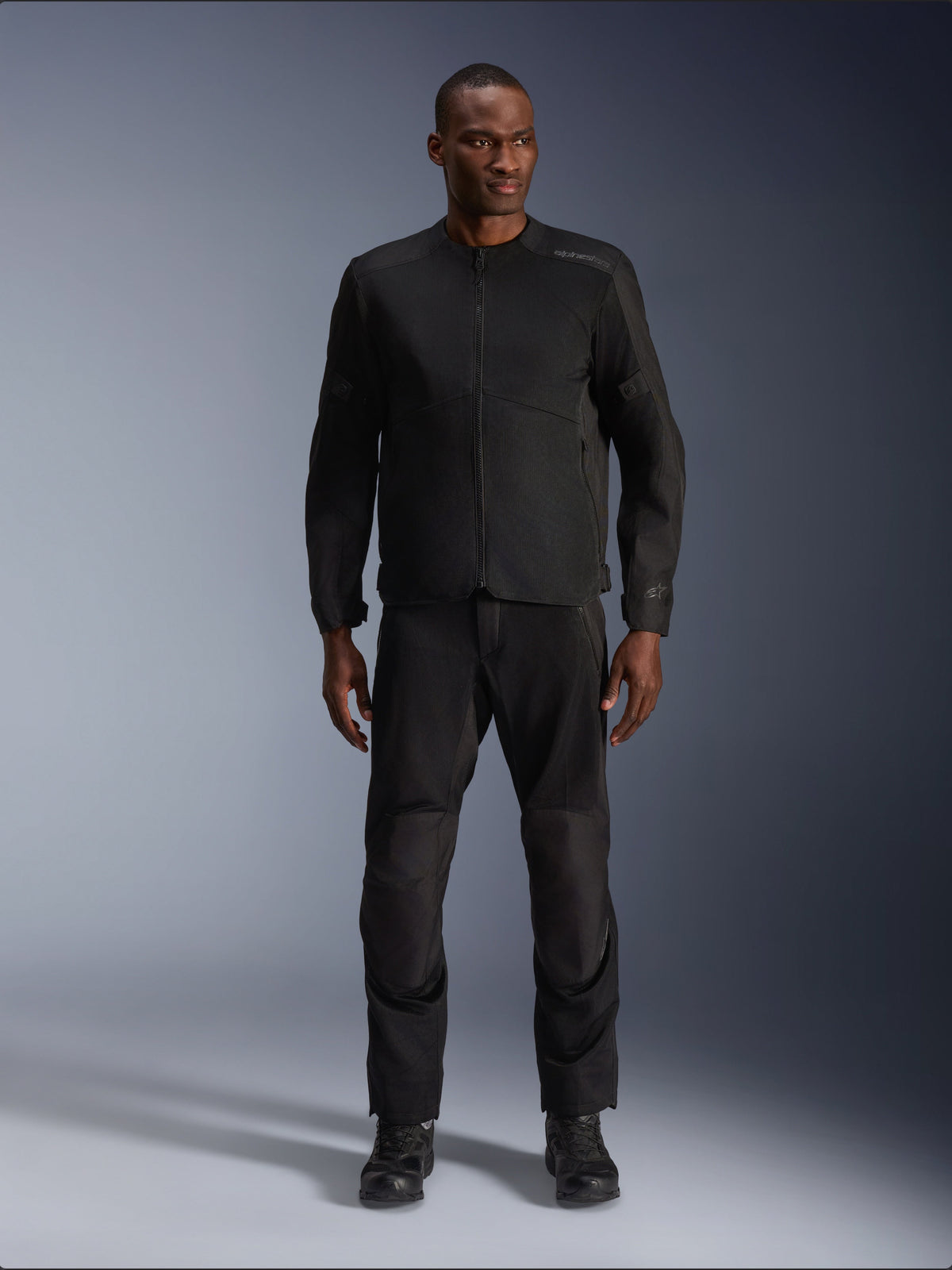 Männliches Model trägt die Alpinestars C-1 Air Jacke in Schwarz, kombiniert mit passender schwarzer Hose und Stiefeln, urbane Motorradausrüstung für Pendler, schlichte Ästhetik mit Mesh-Einsätzen und dezentem Branding.