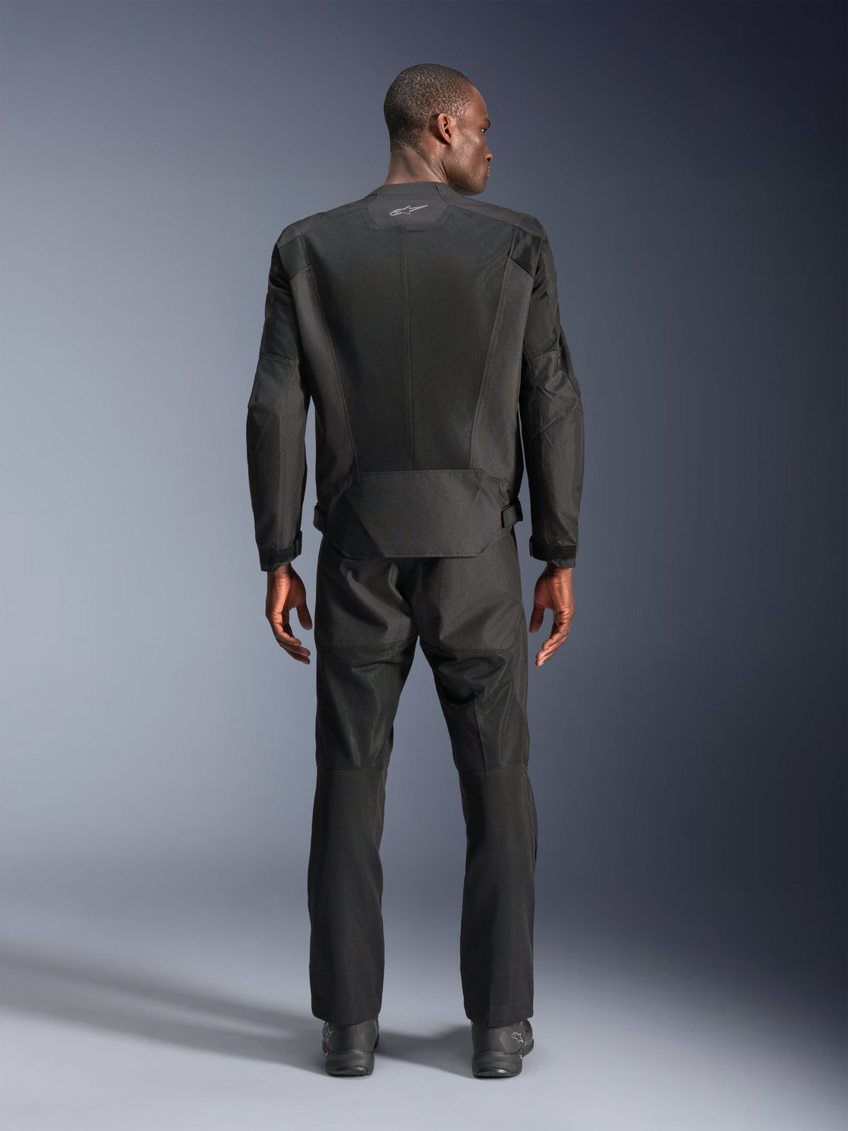 Rückansicht eines Mannes in der Alpinestars C-1 Air Jacke in Schwarz, urbane Motorradjacke mit Tech-Mesh-Einsätzen, passende schwarze Textilhose und Stiefel, schlichte Ästhetik mit reflektierendem Logo am oberen Rücken.