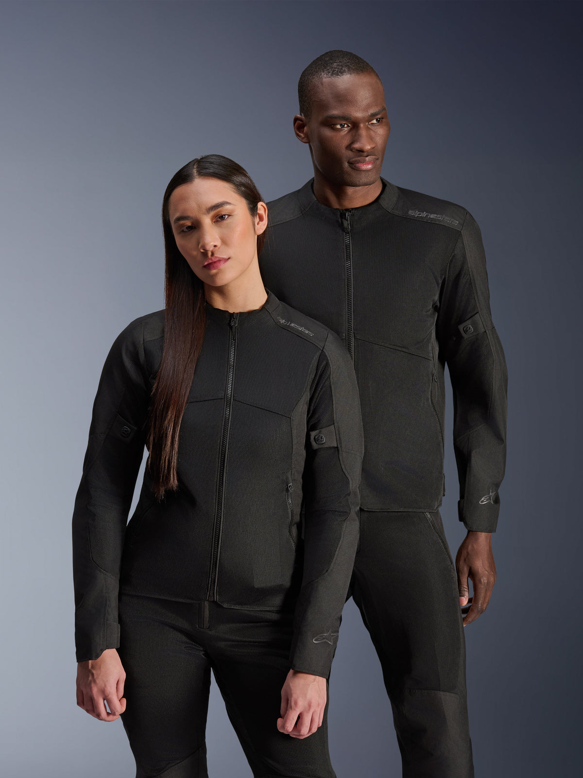 Ein Mann und eine Frau tragen die Alpinestars C-1 Air Jacke und die Stella C-1 Air Jacke, passende schwarze urbane Textil-Motorradjacken mit Mesh-Einsätzen zur Belüftung, kombiniert mit schwarzen Motorradhosen für einen klaren, modernen Look.