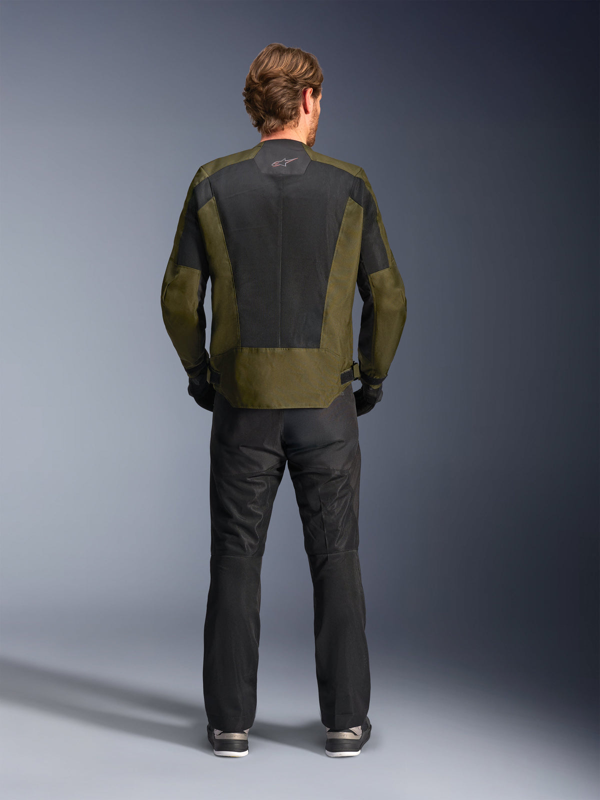 Rückansicht eines Fahrers in der Alpinestars C-1 Air Jacke in Khaki und Schwarz, mit einem großen Tech-Mesh-Rückenpanel zur Belüftung, kombiniert mit einer schwarzen Motorradhose, moderne urbane Straßenbekleidung.