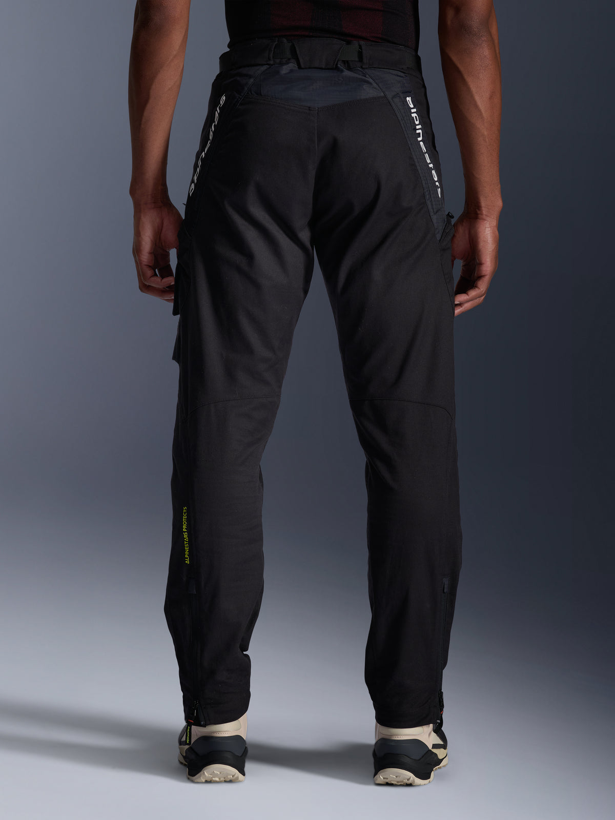 Alpinestars Acteon Hose, Urban-Motorradhose, Schwarz, Rückansicht mit verjüngter Passform, weißes Branding an den Oberschenkeln und gelbes Logo an der unteren Wade, verstellbare Beinabschlüsse mit Reißverschlüssen, kombiniert mit cremefarbenen Sneakern