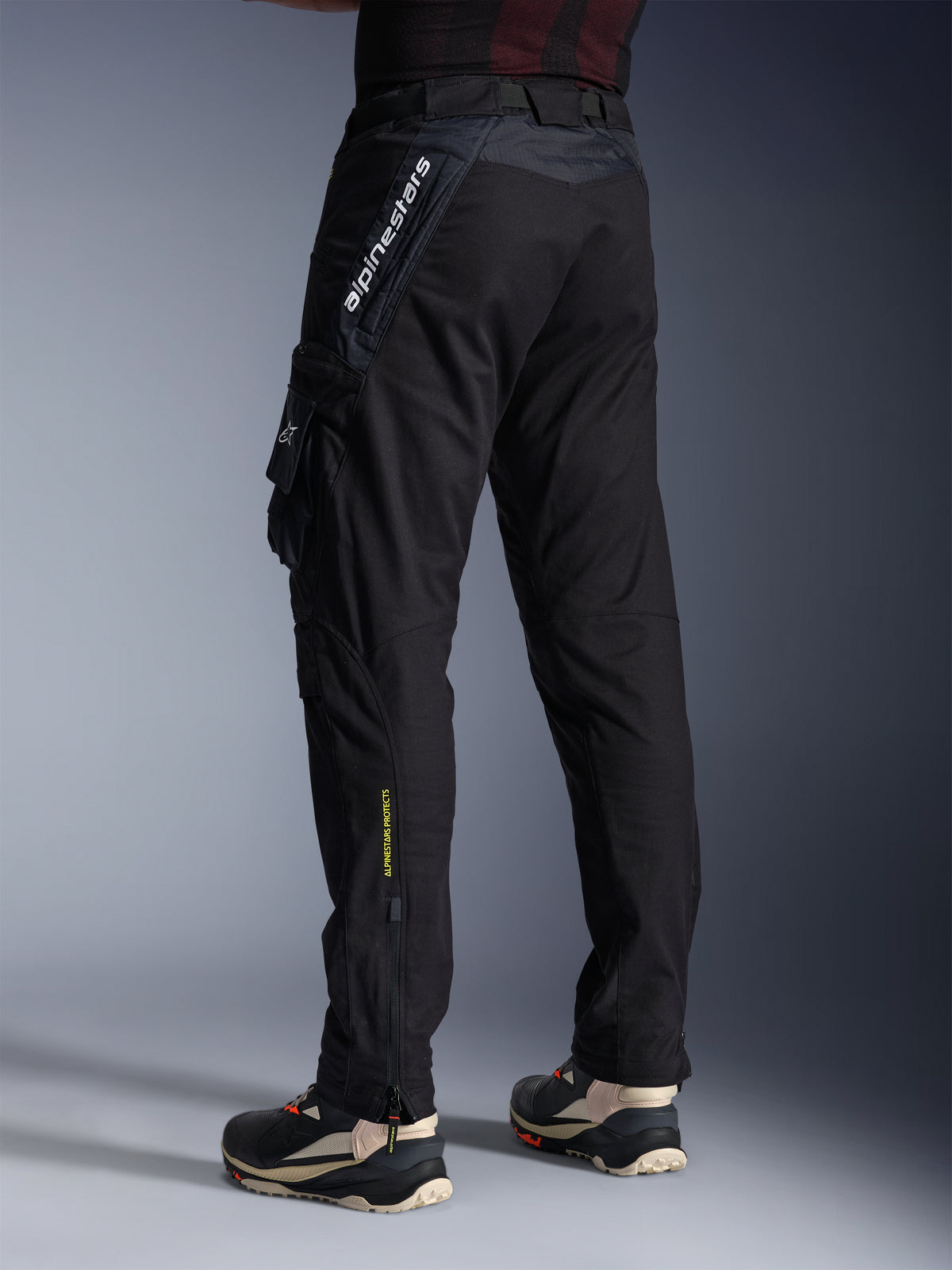 Alpinestars Acteon Hose, schwarze Motorradhose, Rück- und Seitenansicht mit moderner, schmal zulaufender Passform, seitliche Cargotasche mit Logo, vertikaler weißer Alpinestars-Schriftzug am Oberschenkel, gelber „Alpinestars Protects“-Schriftzug am Unterschenkel, moderne urbane Fahrausrüstung