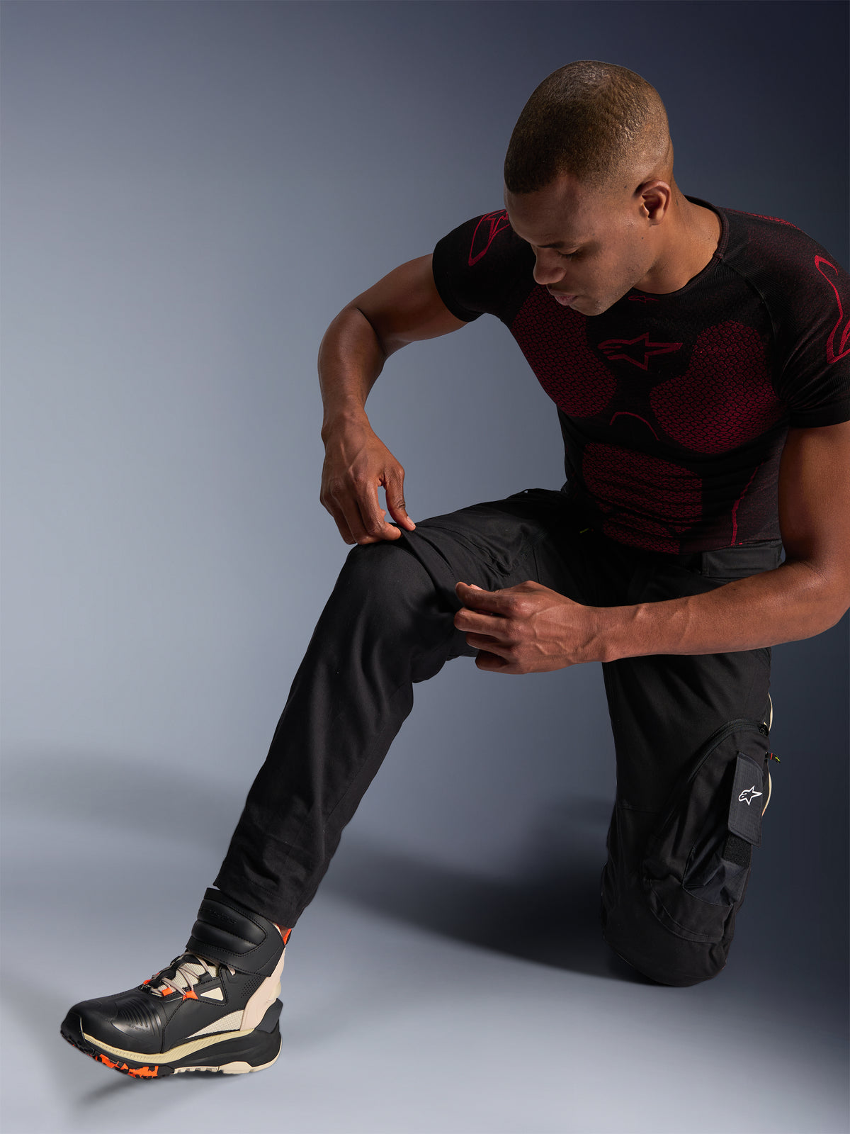 Alpinestars Acteon Hose, schwarze urbane Motorradhose, getragen von einem knienden Mann, der den Kniebereich anpasst, mit Cargotaschen und einer schmal zulaufenden Passform, kombiniert mit einem schwarz-roten Alpinestars Base Layer und Motorradschuhen