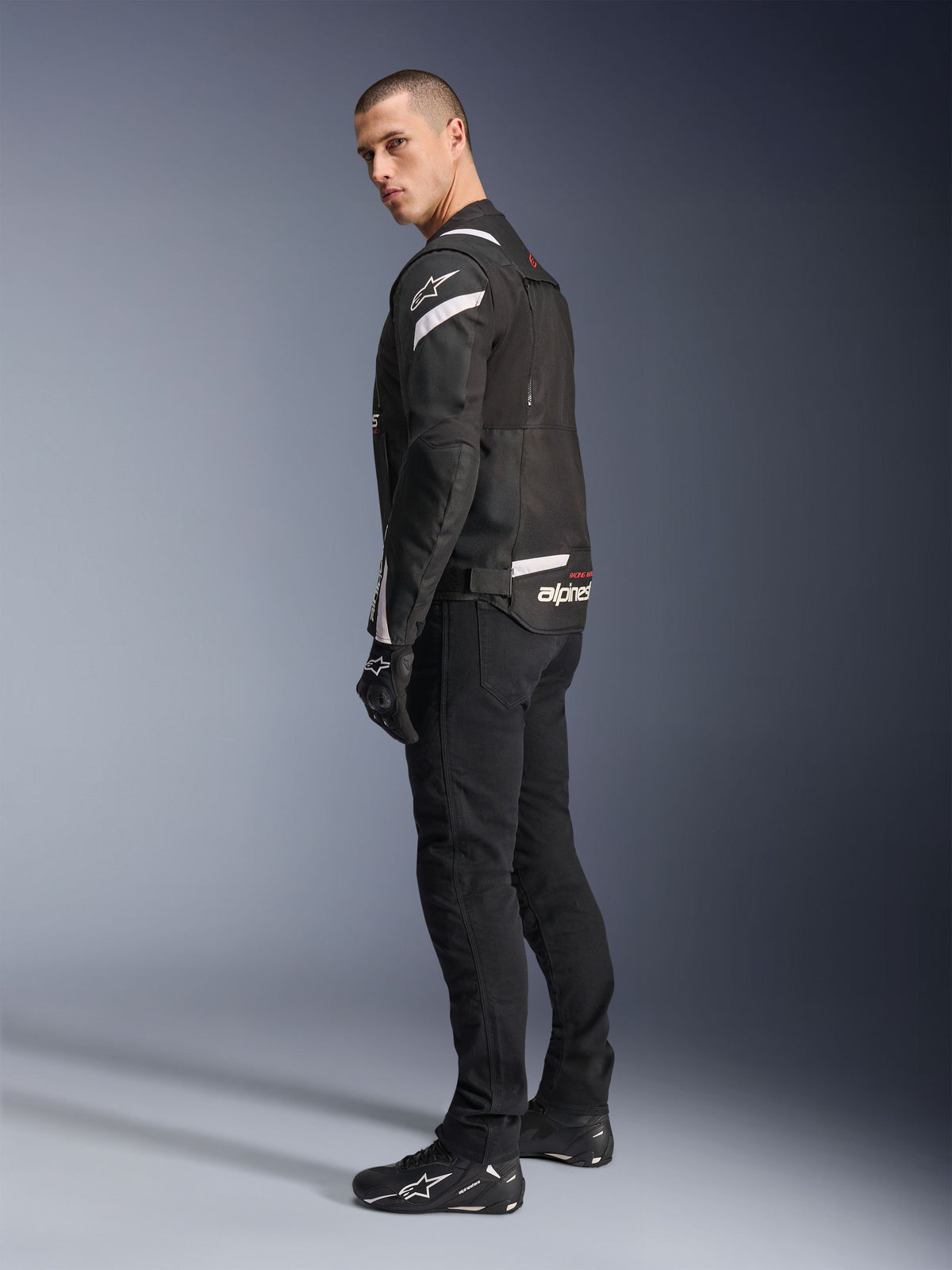 Alpinestars T-Stunt Air Jacke, Schwarz und Weiß, modulare Motorradjacke mit Tech-Mesh-Einsätzen, Rückansicht im Profil an einem Modell, getragen mit schwarzer Fahrhose und Schuhen, mit weißen Akzenten und Branding auf den Schultern und dem Rücken.