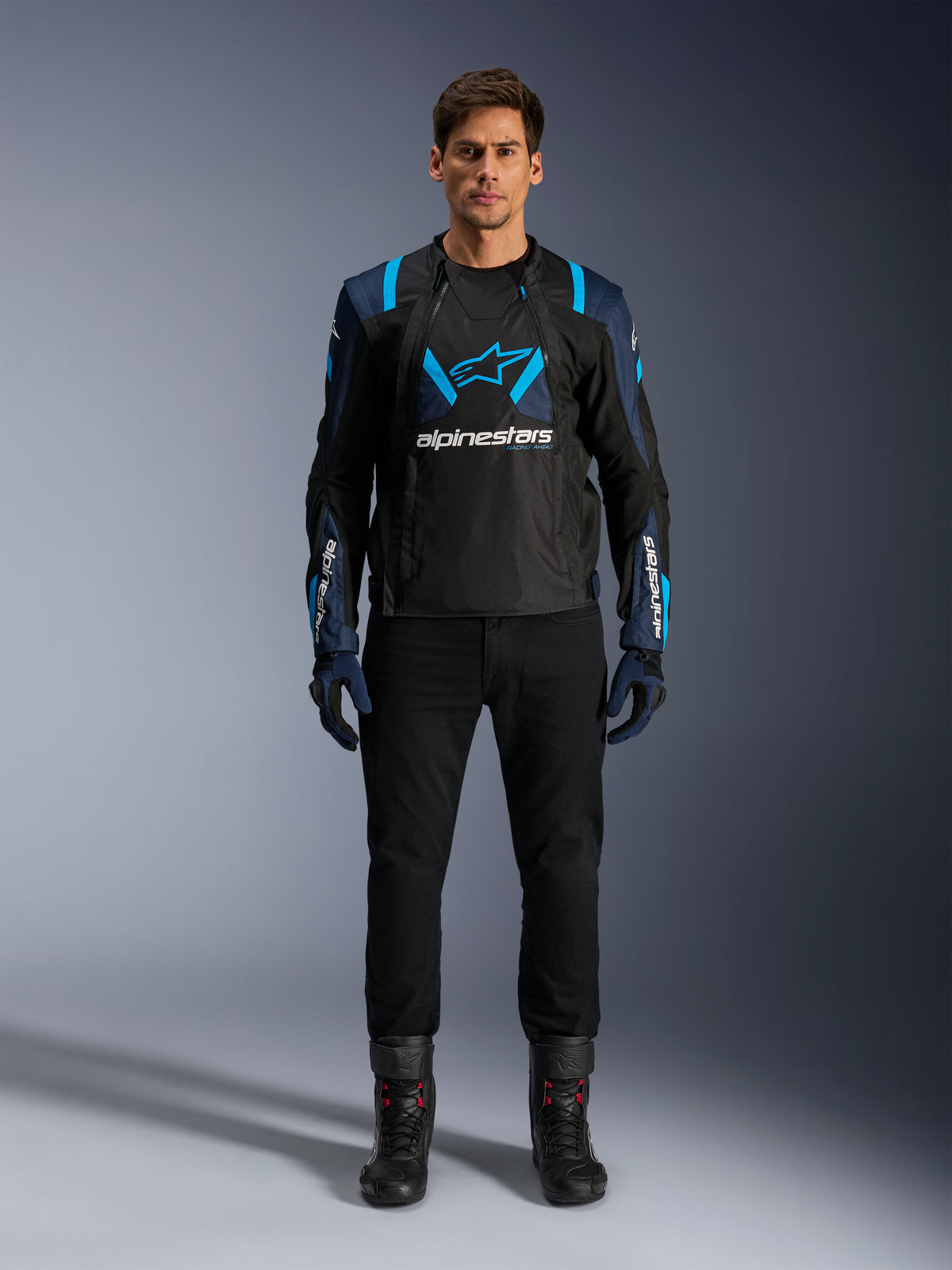 Alpinestars T-Stunt Air Jacke, Motorradjacke, Schwarz mit dunkelblauen und hellblauen Akzenten, mit einem großen weißen Logo auf der Brust, sportlich geschnittene Mesh-Konstruktion für optimale Belüftung, getragen mit schwarzer Motorradhose und Stiefeln für eine elegante Road-Riding-Ästhetik.