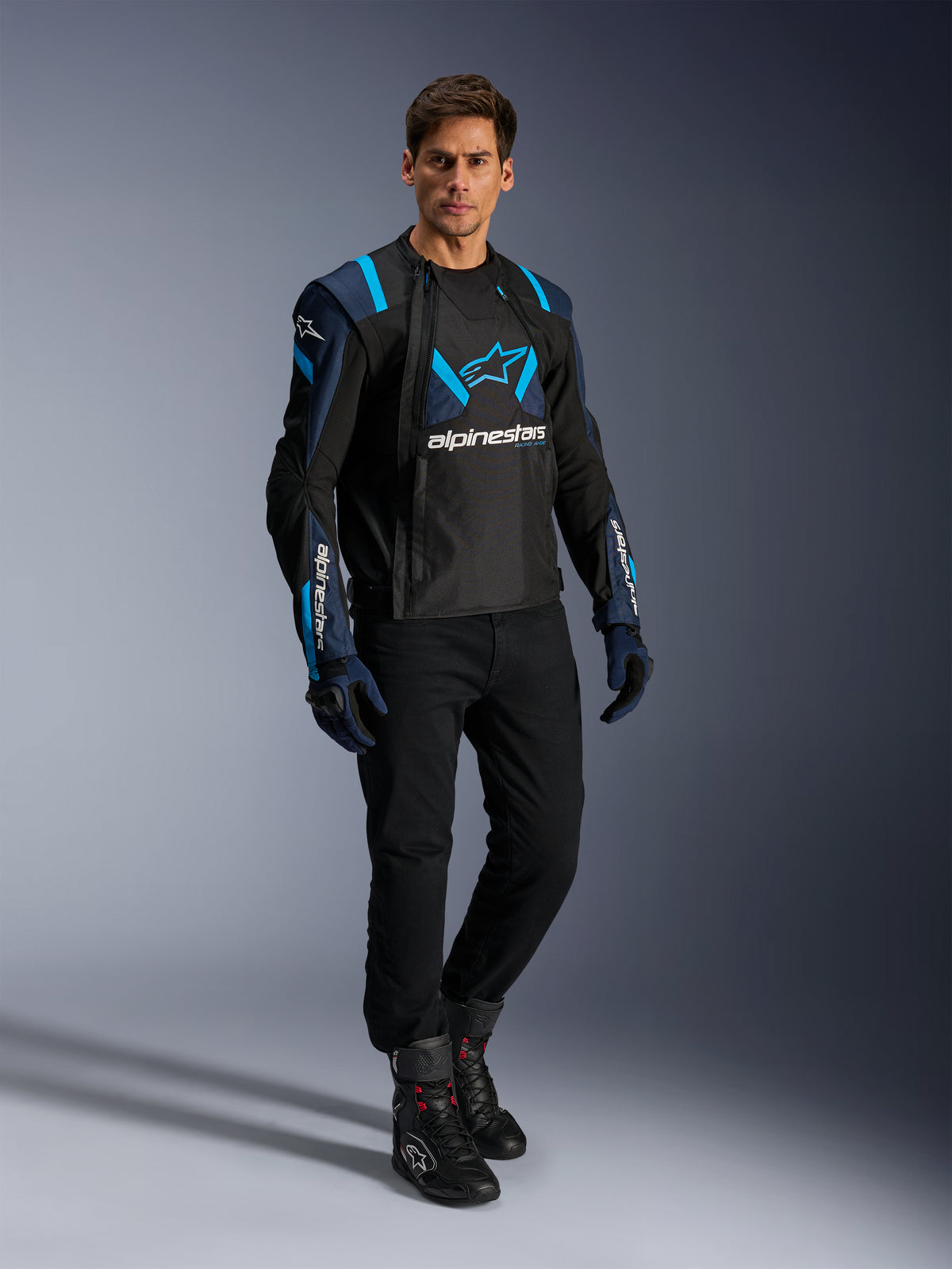 Männliches Model in Alpinestars T-Stunt Air Jacke, Schwarz, Dunkelblau und Blithe Blue, mit markanten Mesh-Einsätzen für Luftzirkulation, kombiniert mit schwarzer Hose und Motorradhandschuhen, sportlich geschnittene Motorradjacke für sommerliche Straßenfahrten