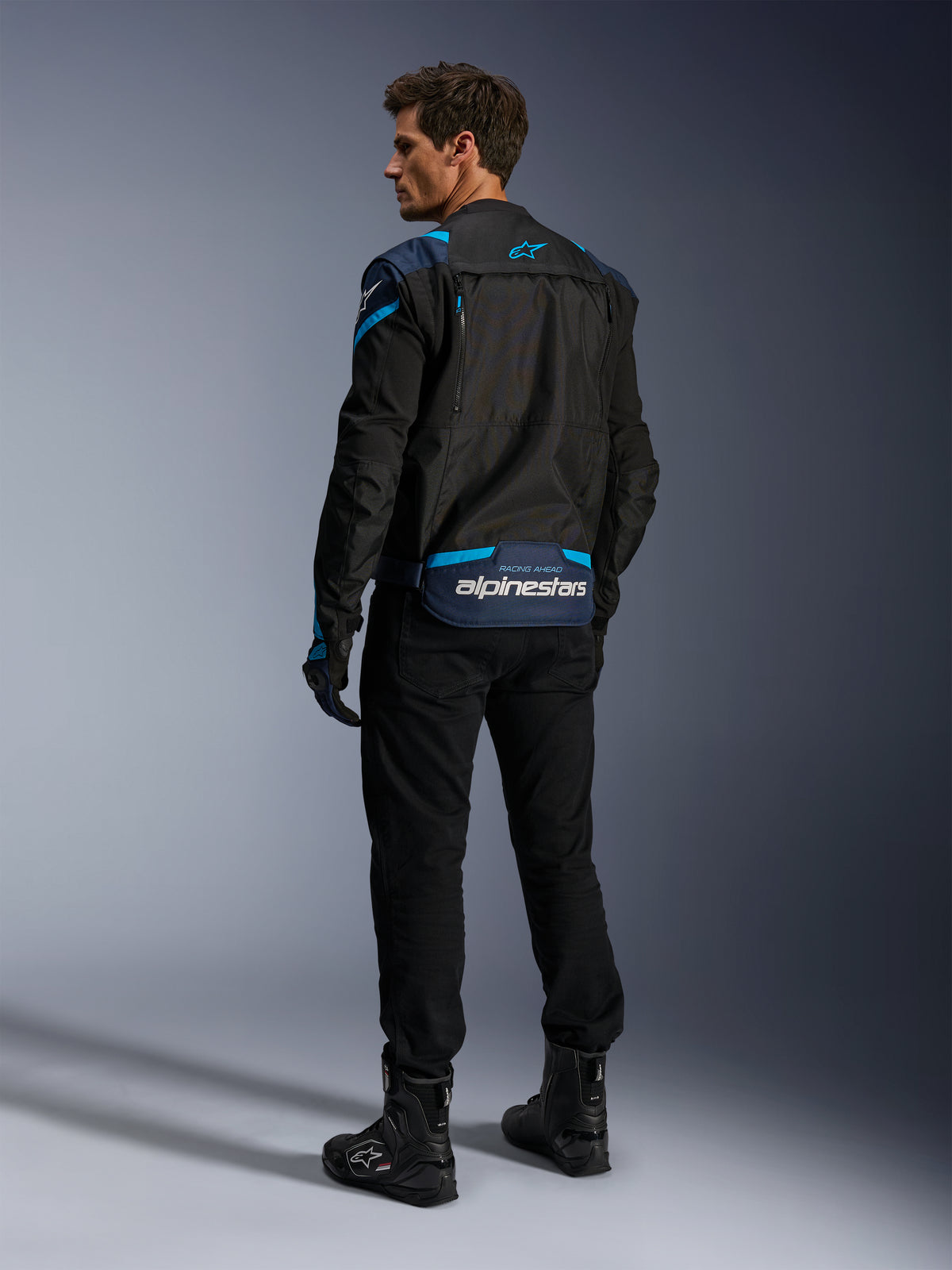 Alpinestars T-Stunt Air Jacke, Motorradjacke, Schwarz mit dunkelblauen und hellblauen Akzenten, Rückansicht mit modularen Mesh-Einsätzen und Branding am unteren Rücken, getragen von einem Modell mit schwarzer Motorradhose und Stiefeln.