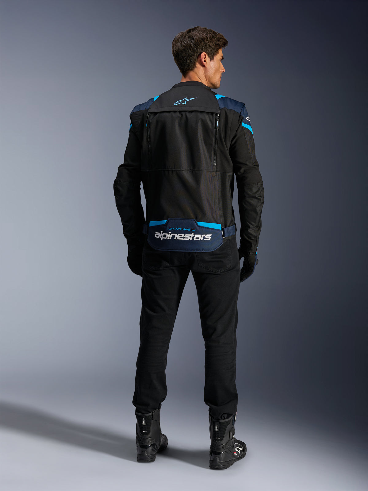 Alpinestars T-Stunt Air Jacke, modulare Textil-Motorradjacke, Schwarz, Dunkelblau und Hellblau, Rückansicht zeigt das Staufach für die Ärmel im unteren Rückenbereich mit weißem Alpinestars-Branding, getragen mit schwarzer Hose und Stiefeln.