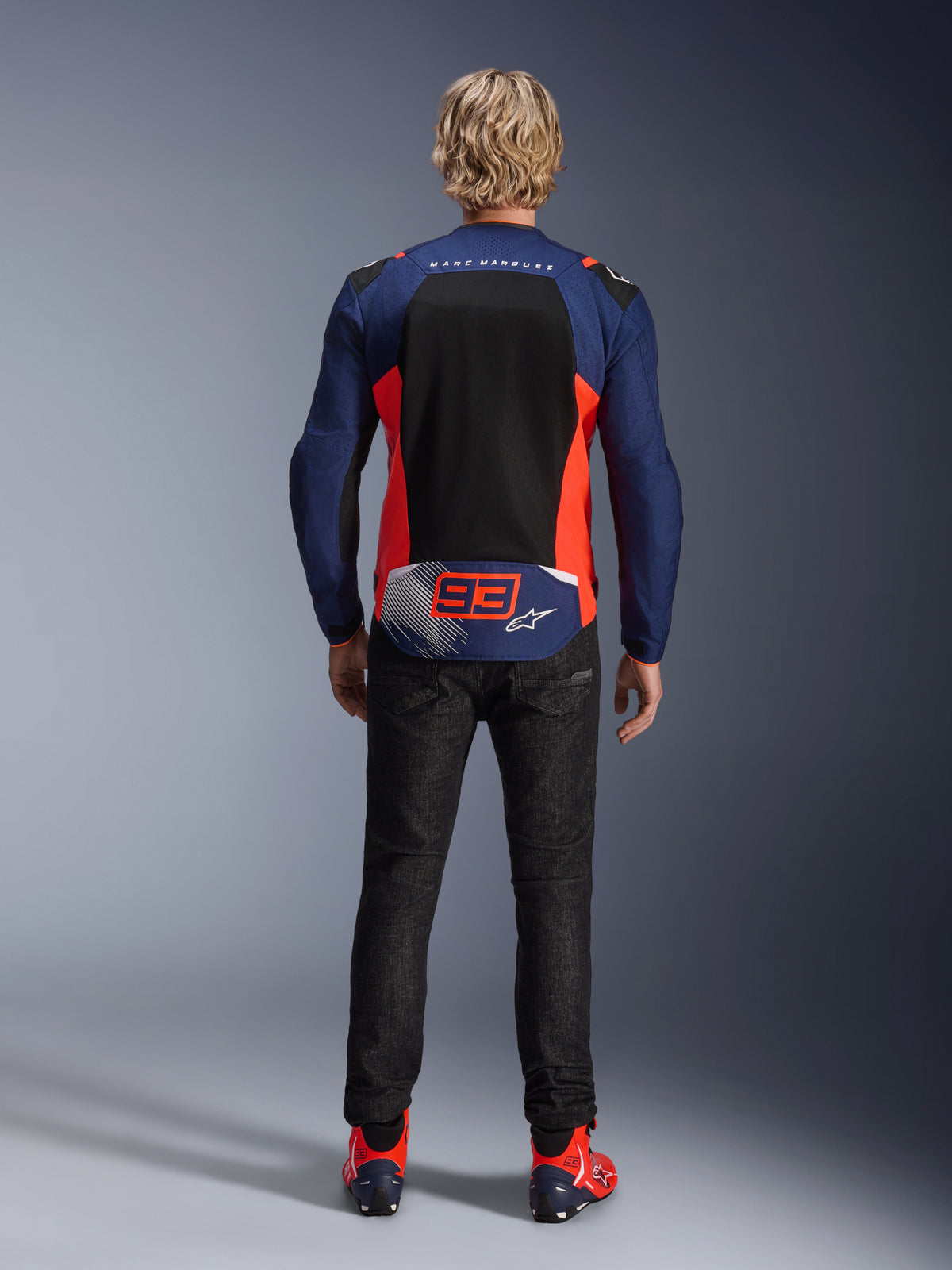 Rear view of a model wearing the MM93 T-SPS Air V2 Jacket in Blue Navy with Red Fluo and Black accents, featuring a large mesh back panel and '93' Marc Marquez branding, paired with dark denim pants and matching riding boots