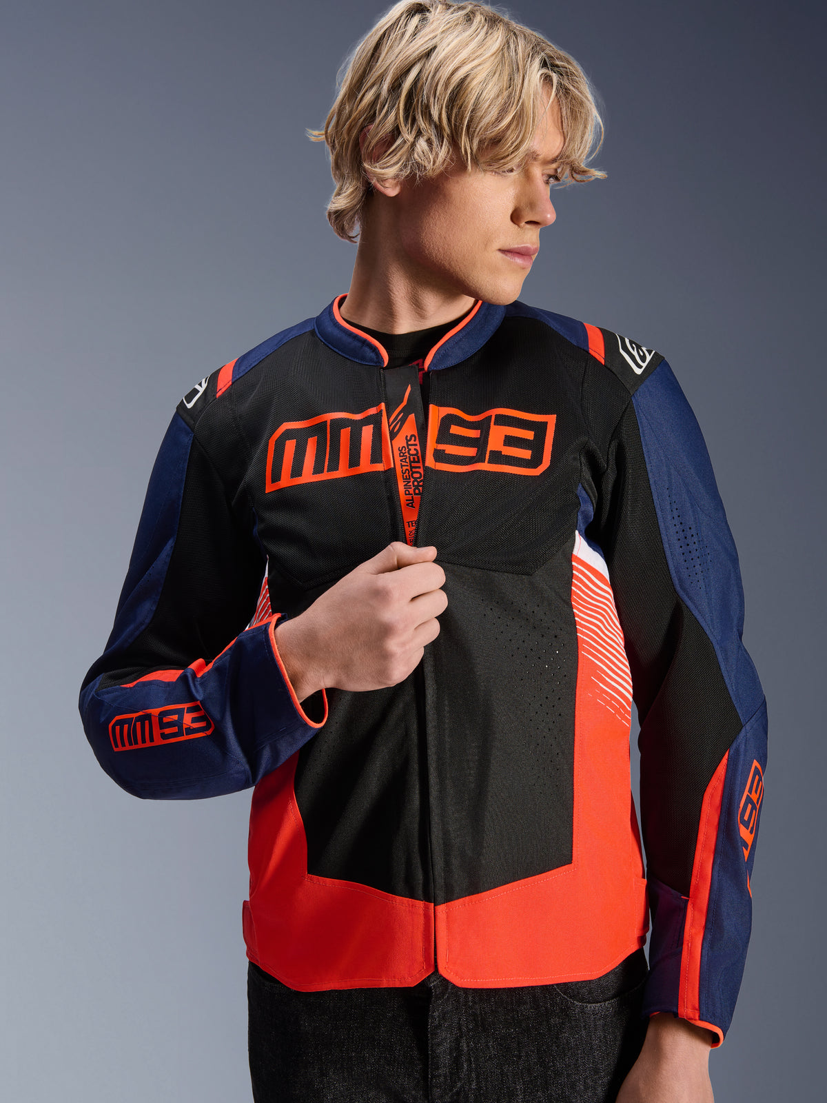 Alpinestars MM93 T-SPS Air V2 Jacket, Navy Blue, Black and Fluorescent Red textile motorcycle jacket, worn by a male model adjusting the zipper, featuring bold Marc Márquez MM93 branding on the chest and sleeves, with ventilated mesh panels for summer road riding.