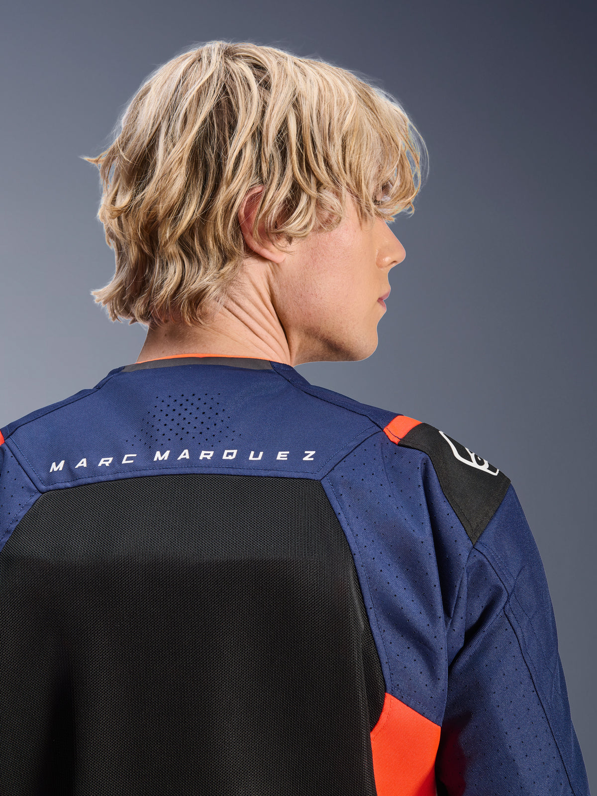 Alpinestars MM93 T-SPS Air V2 Jacket, Blue Navy and Black with Red Fluo accents, close-up of the upper back featuring 'MARC MARQUEZ' branding, breathable mesh panels and perforated textile construction, worn by a blonde model.