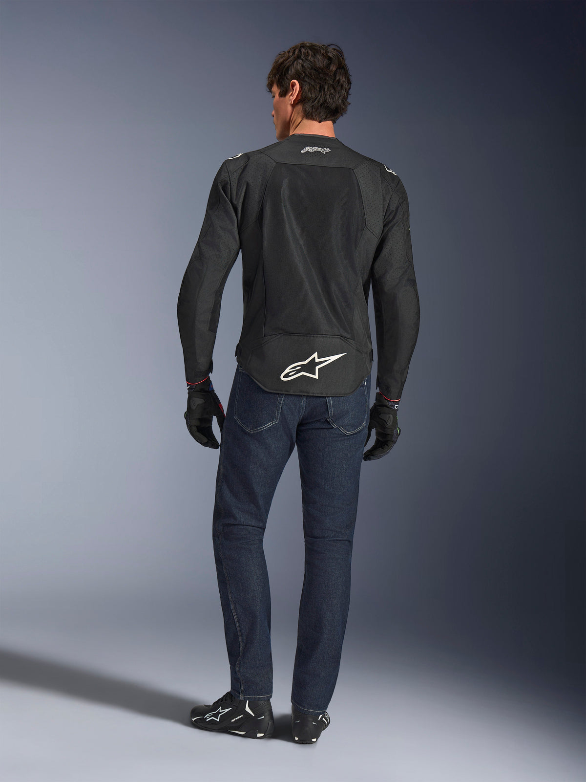 Alpinestars FQ20 T-SPS Air V2 Monster Jacke, Schwarz, Rückansicht an einem Model in dunkelblauen Jeans und schwarzen Motorradstiefeln, mit großem weißem Logo am unteren Rücken und atmungsaktiven Mesh-Einsätzen, urbane Sport-Motorradausrüstung