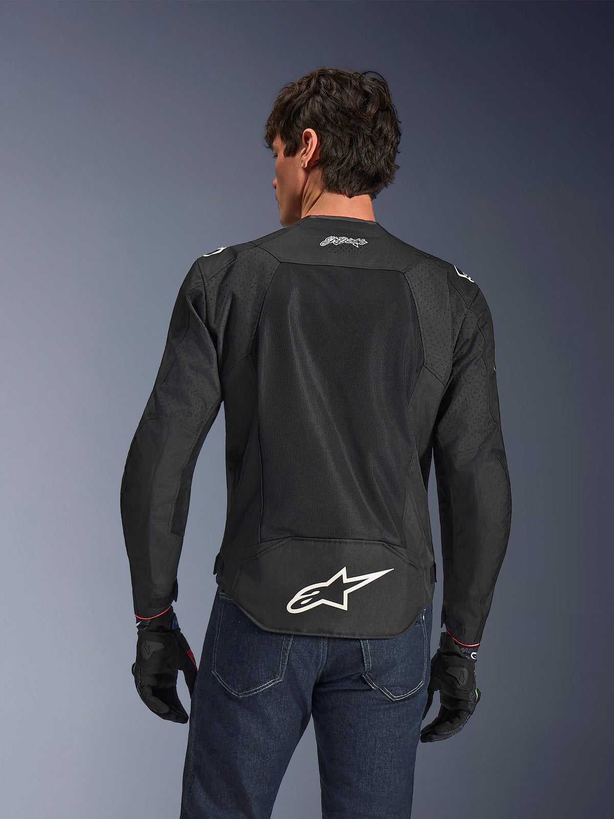 Alpinestars FQ20 T-SPS Air V2 Monster Jacke, Schwarz, Rückansicht mit großen Mesh-Belüftungseinsätzen und weißem Alpinestars-Logo am unteren Rücken, getragen mit blauen Jeans und schwarzen Motorradhandschuhen, urbaner Sport-Fahrstil.