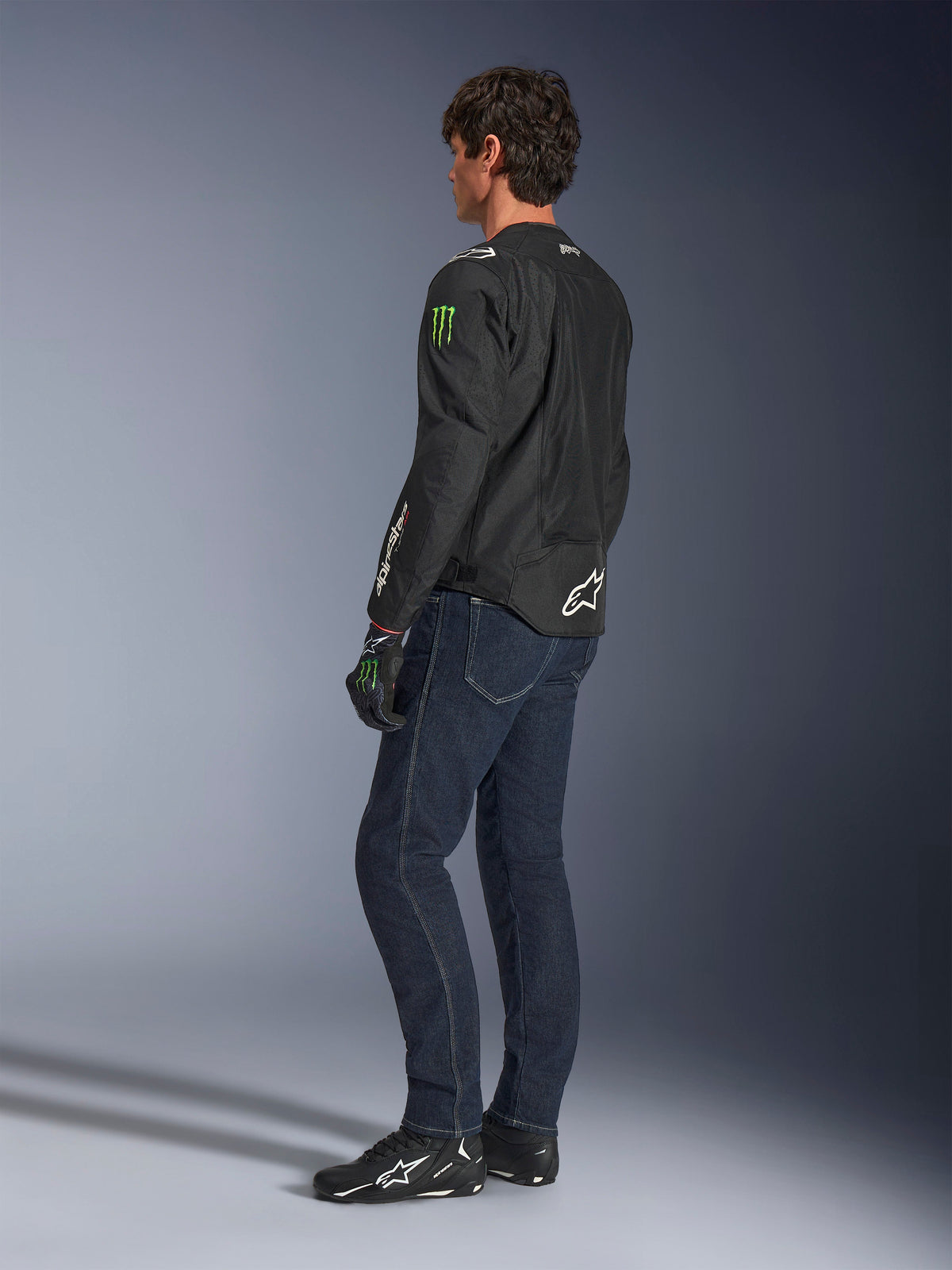 Alpinestars FQ20 T-SPS Air V2 Monster Jacke, schwarze Textil-Motorradjacke mit grünen Monster Energy-Logos auf den Ärmeln und weißem Alpinestars-Branding, getragen von einem Modell in der Dreiviertel-Rückansicht mit dunkelblauen Jeans und schwarzen Motorradschuhen, urbaner Sport-Stil.