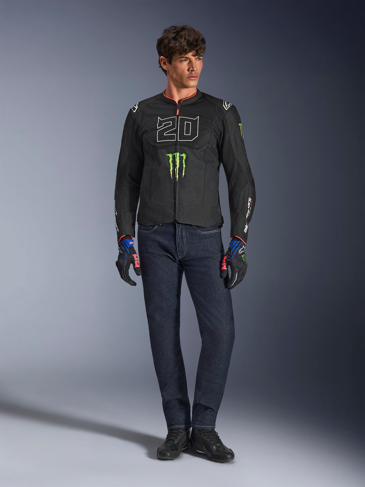 Männlicher Fahrer trägt die Alpinestars FQ20 T-SPS Air V2 Monster Jacke, schwarz mit grünem Monster Energy Logo und weißer „20“-Grafik auf der Brust, kombiniert mit dunkelblauen Jeans und technischen Fahrhandschuhen, urbanes Sport-Motorrad-Outfit.