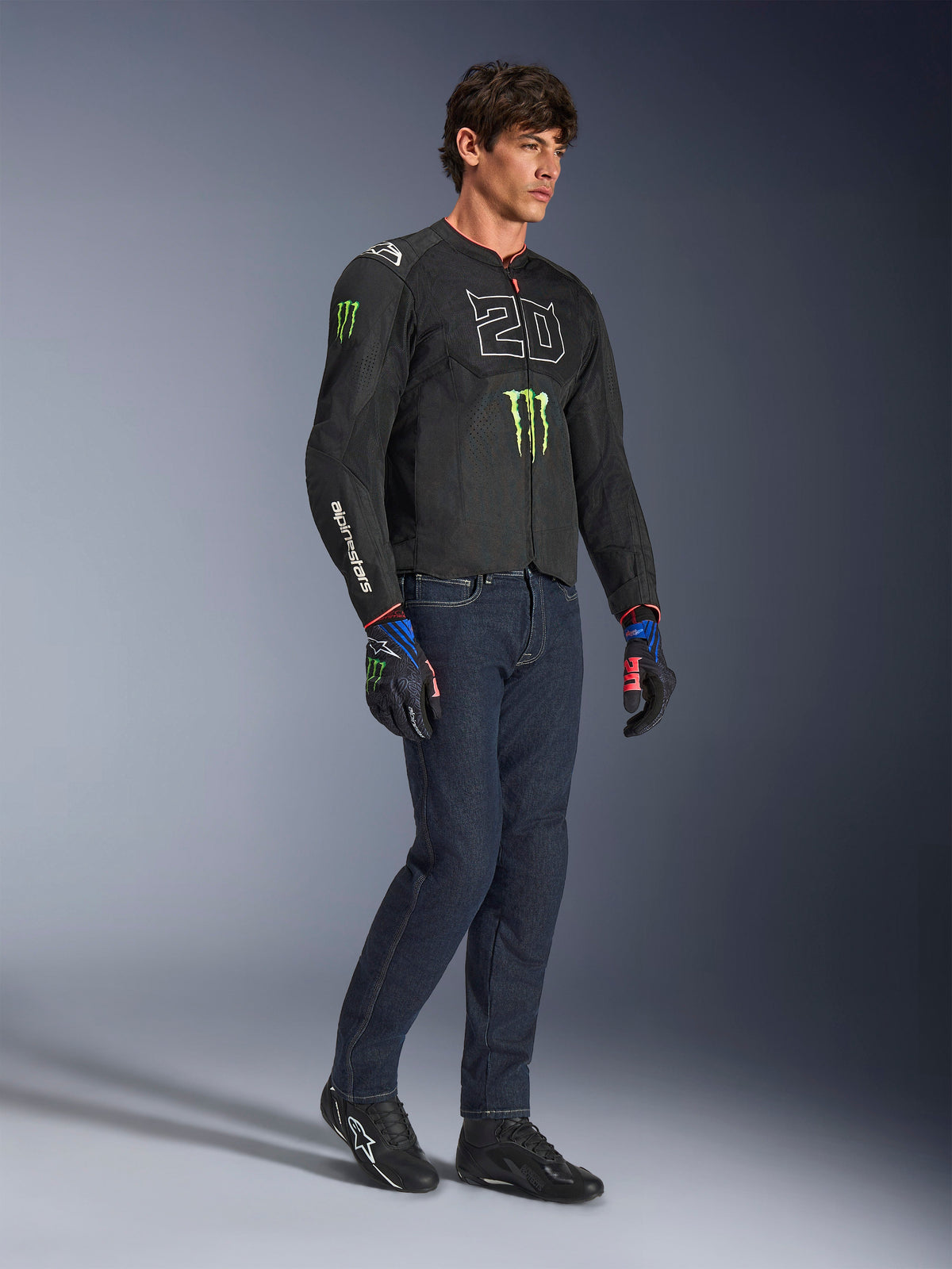 Männlicher Fahrer trägt die Alpinestars FQ20 T-SPS Air V2 Monster Jacke in Schwarz, mit dem „20“-Logo und der Monster Energy-Kralle auf der Brust, kombiniert mit dunkler Motorradjeans, schwarzen Motorradschuhen und passenden FQ20 Handschuhen, Ganzkörper-Studioaufnahme.