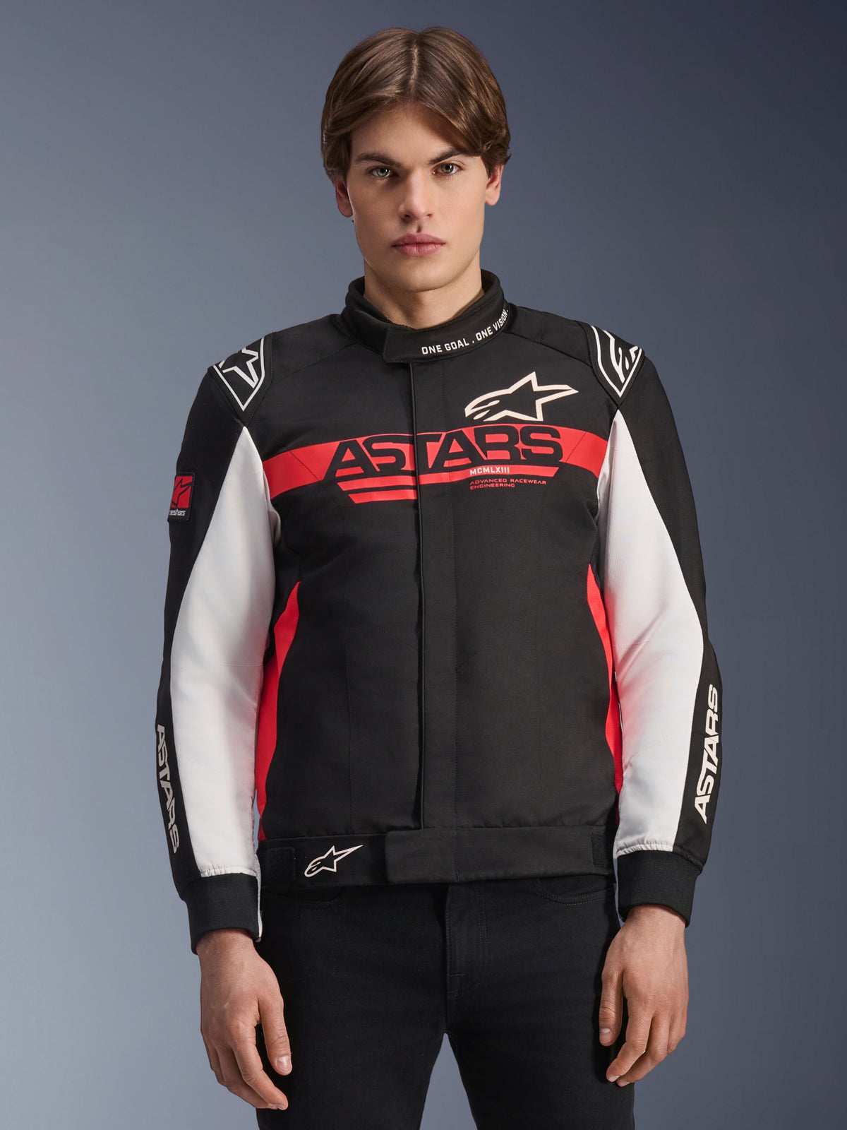 Alpinestars Monza Sport Jacke, Textil-Motorradjacke, Schwarz mit hellroten und weißen Akzenten, markantes ASTARS-Logo auf der Brust, 'ONE GOAL. ONE VISION.' Schriftzug am Kragen, weiße Ärmelpartien und Stern-Logos, getragen von einem Model vor einem Hintergrundverlauf