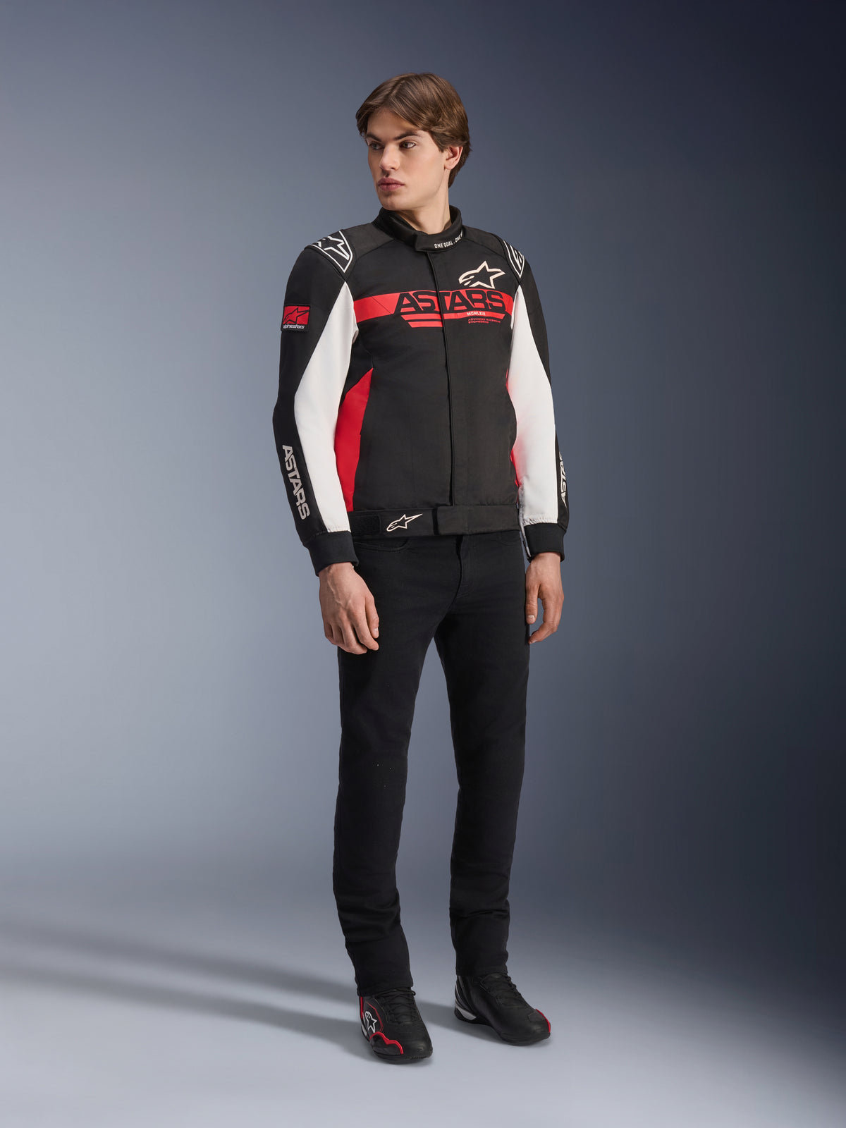 Alpinestars Monza Sport Jacke, Schwarz, Hellrot und Weiß, Textil-Motorradjacke, vom Rennsport inspiriertes Design mit ASTARS-Branding auf der Brust und den Ärmeln, getragen von einem Modell mit schwarzer Hose und Motorradschuhen, lässige Sport-Fahrerausrüstung