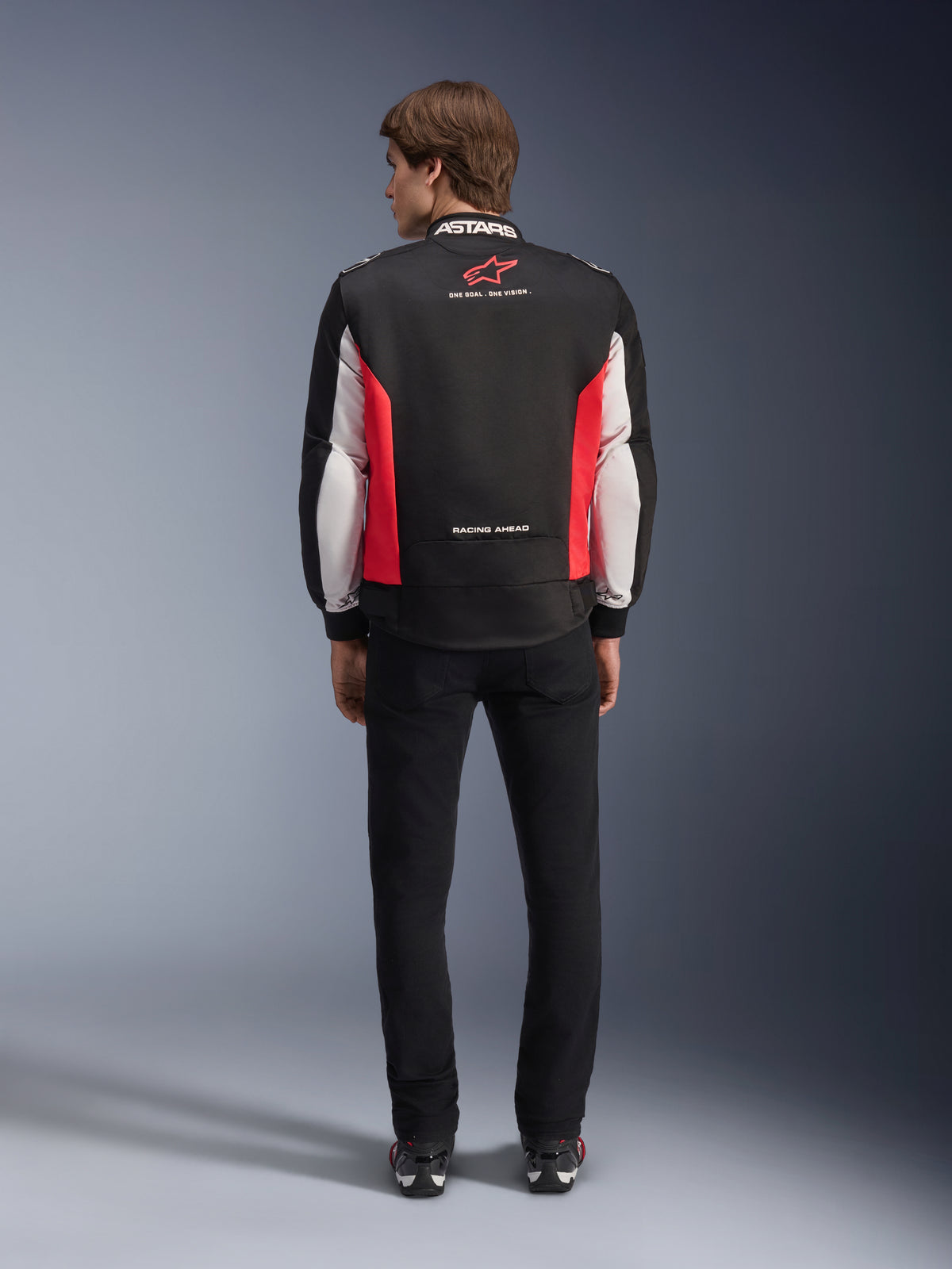 Alpinestars Monza Sport Jacke, Textil-Motorradjacke in Schwarz, leuchtendem Rot und Weiß, Rückansicht an einem Model, mit rotem Stern-Logo und „ASTARS“-Schriftzug am Kragen sowie Slogans, getragen mit schwarzer Hose und Motorradstiefeln.